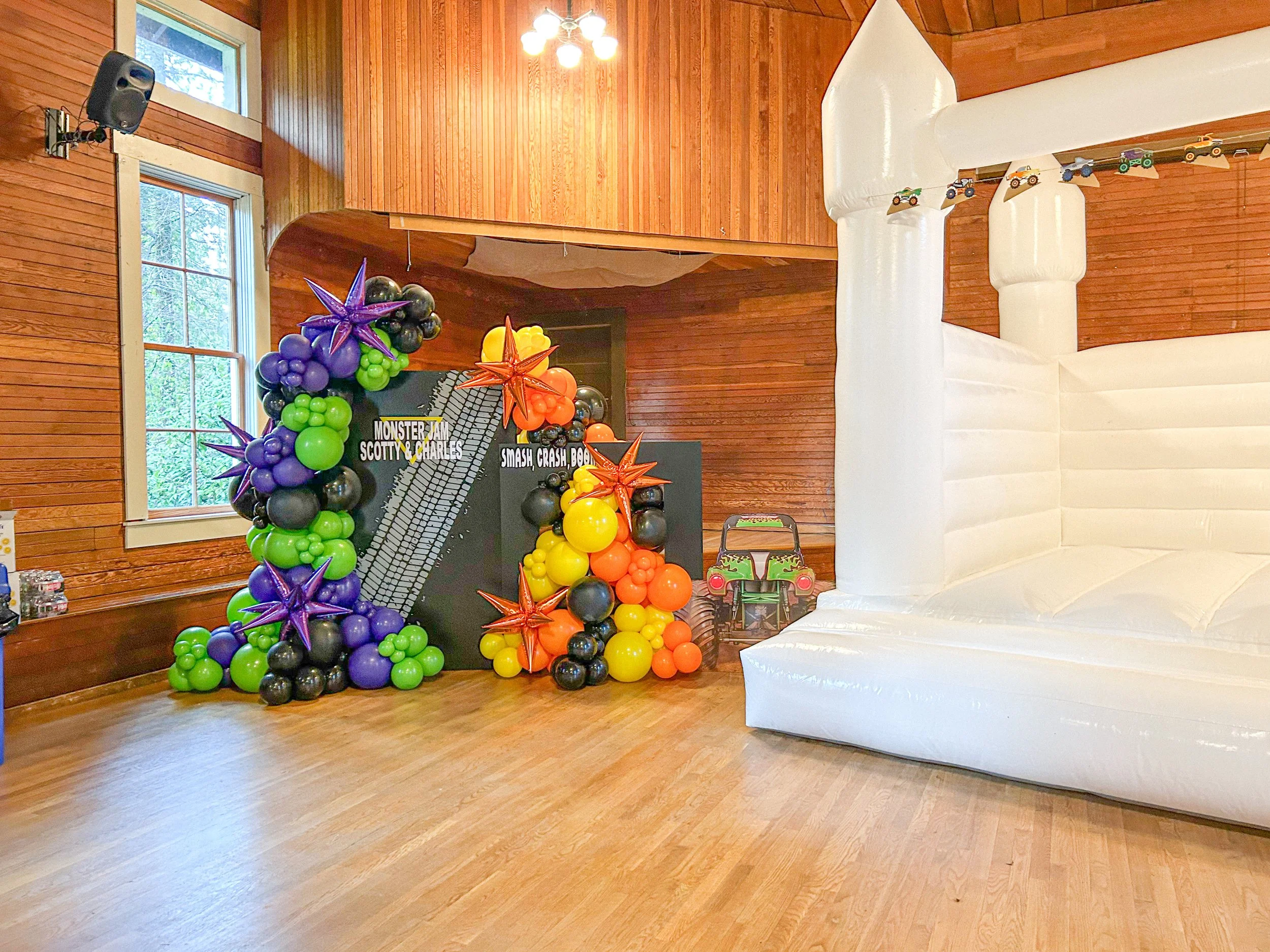 Colorful balloon decorations and an inflatable white bounce house in a wooden room, set up for a children's party with a game area labeled "Monster Jam Scotty & Charles" and featuring small vehicles.