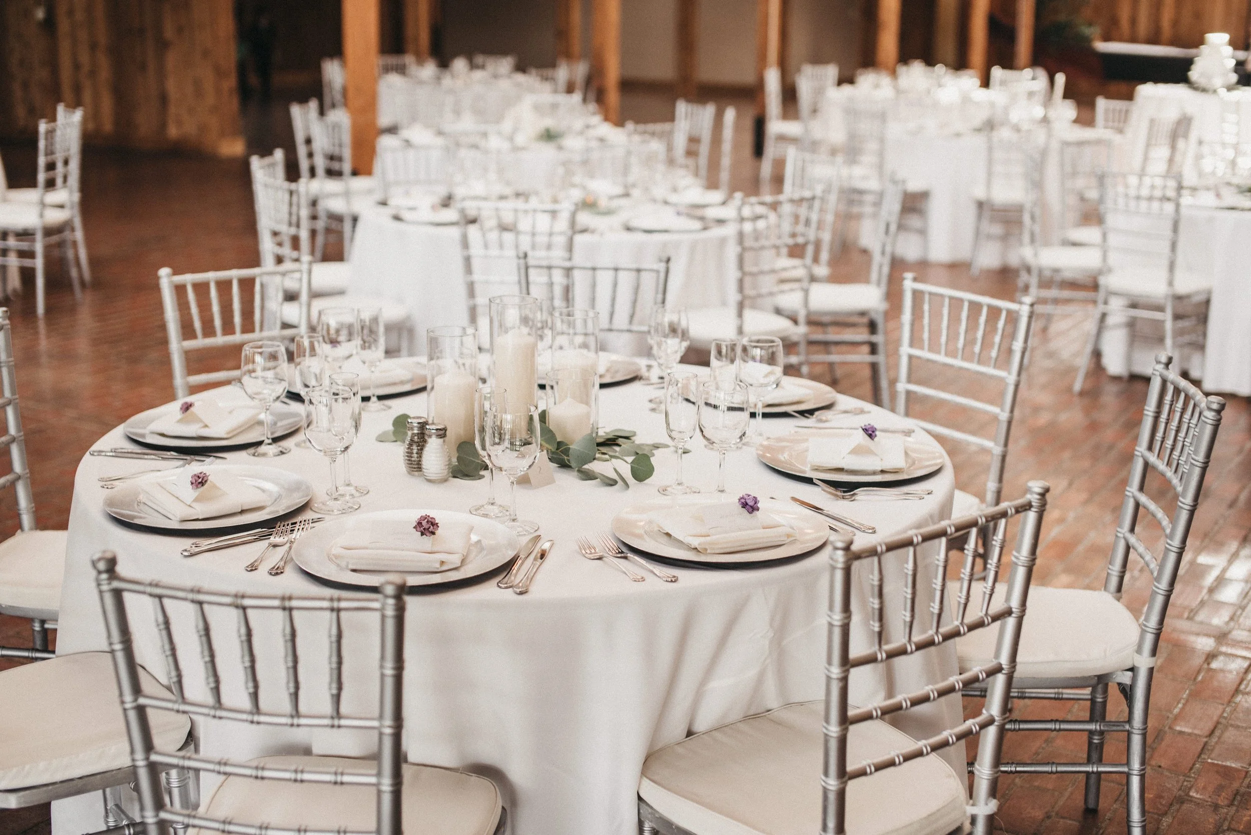 Wedding reception tables decorated with white tablecloths, candles, eucalyptus, and purple flowers, set with plates, silverware, and glassware, arranged in a rustic venue with brick floors and wooden accents.