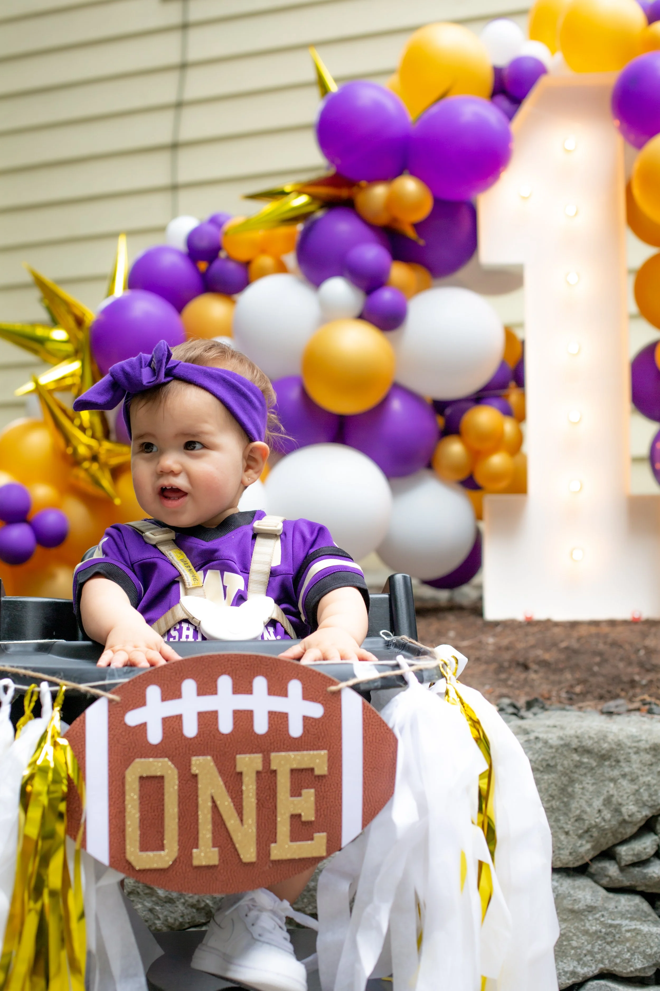 A young girl dressed in purple, sitting in a decorated stroller, celebrating her first birthday with purple, white, and gold balloons in the background and a large lit-up number one.
