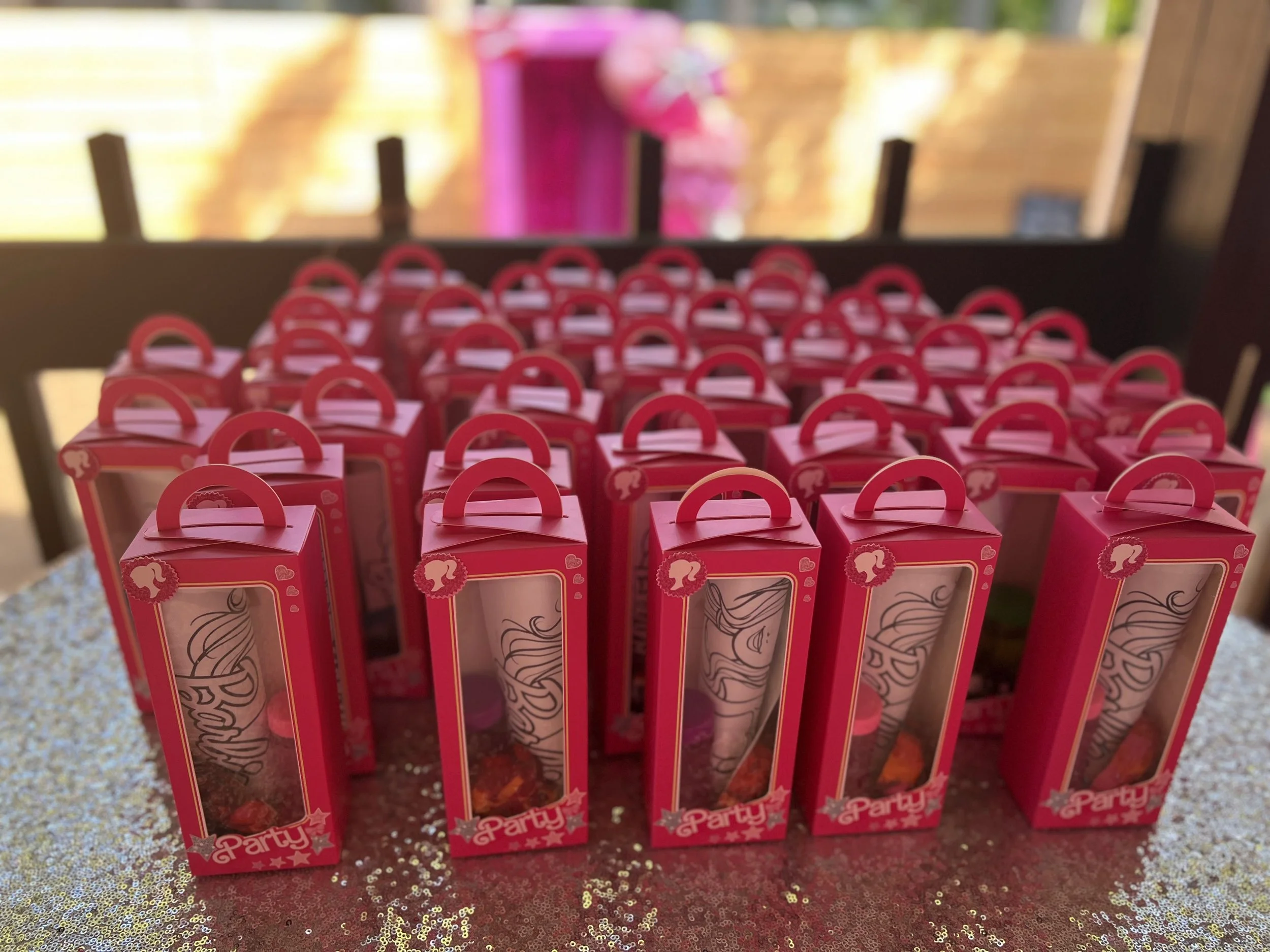 Party favor boxes with pink packaging and transparent windows, decorated with unicorn and cupcake icons, arranged on a glittery tablecloth at a celebration.