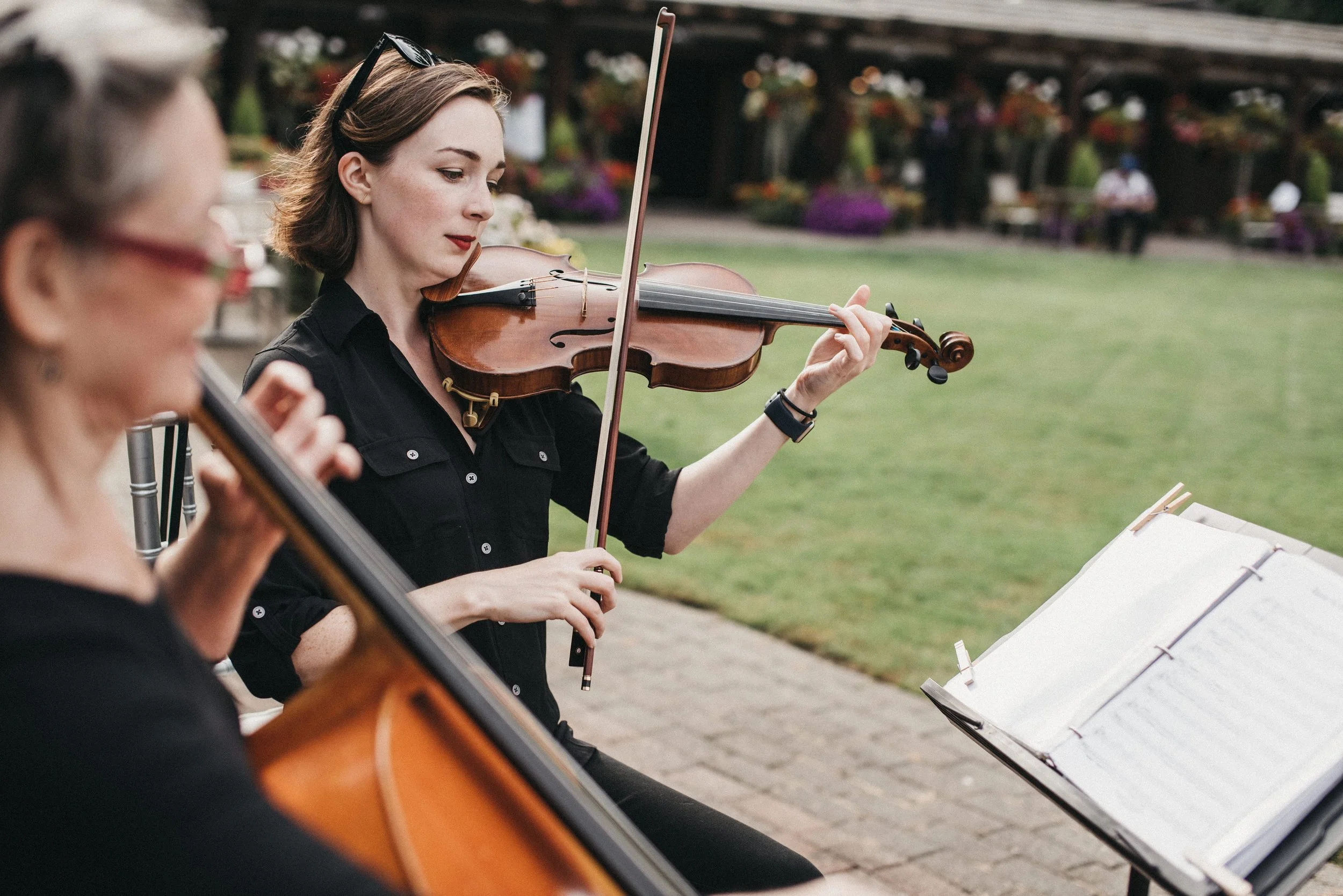 Two women playing violins outdoors, with a music stand in front of them and a background of colorful flowers and people.