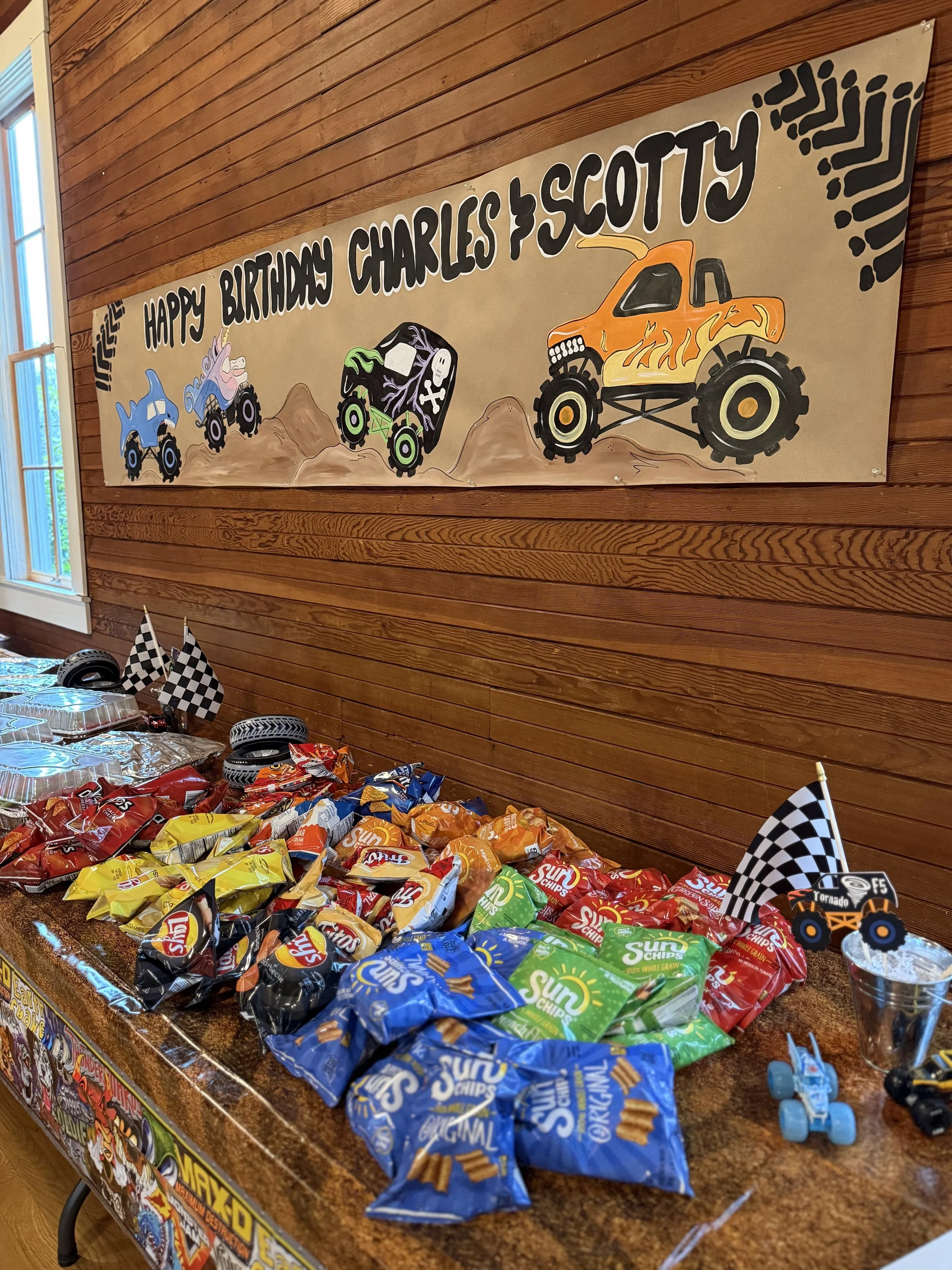 Party table with various bags of Lay's Sun Chips and other snacks, decorated with checkered flags and toy monster trucks, under a birthday banner with Monster Truck theme.