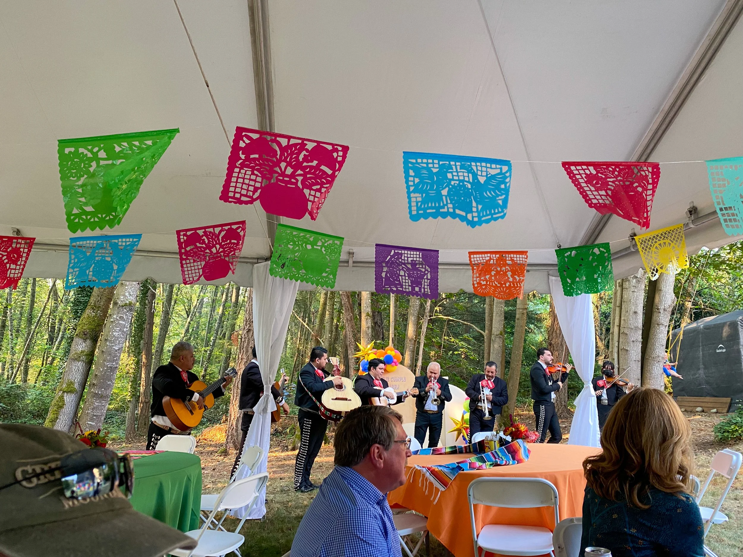 Mexican celebration with a live mariachi band playing in a forested outdoor setting under a colorful papel picado banner.