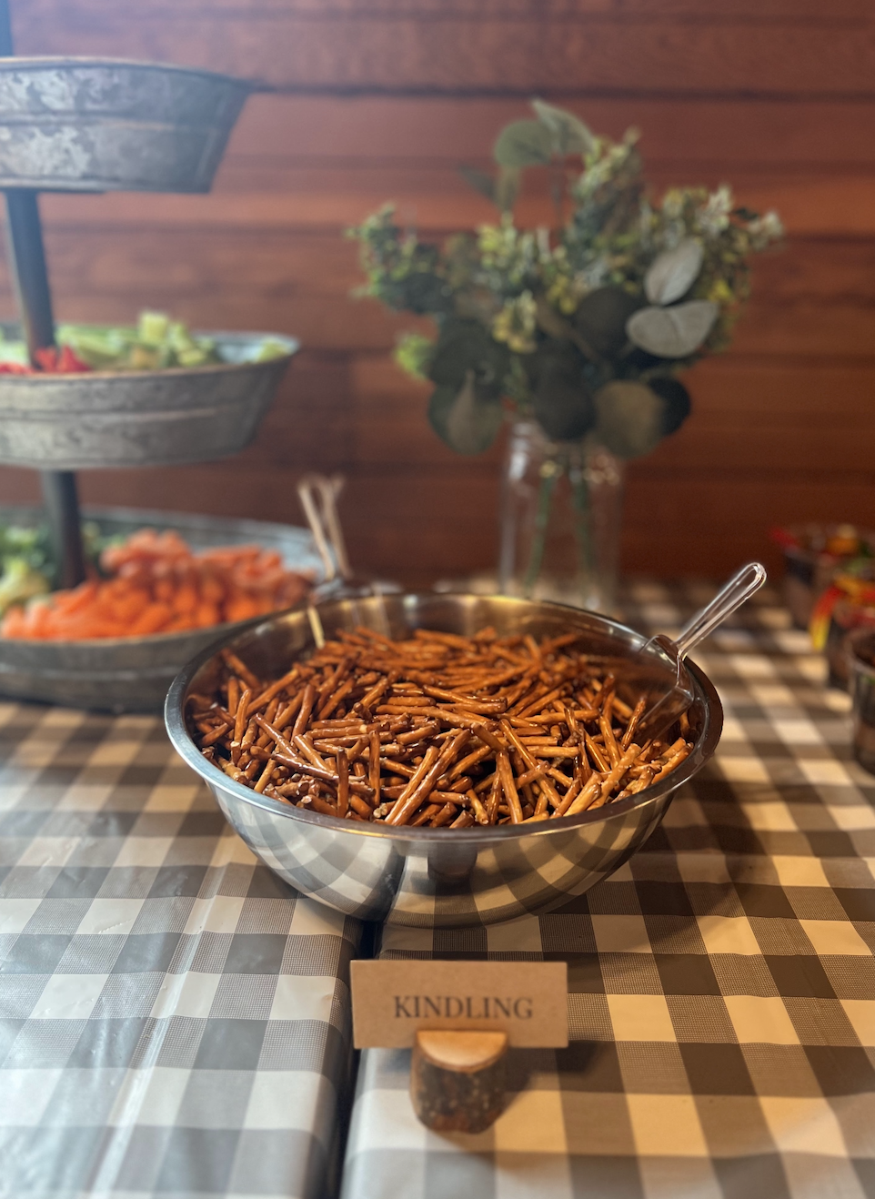 A large metal bowl filled with cooked, seasoned pasta or noodles on a checkered tablecloth, with a small sign reading 'KINDLING' in front of it. In the background, there are layered trays of sliced vegetables, a glass jar with a floral arrangement, a