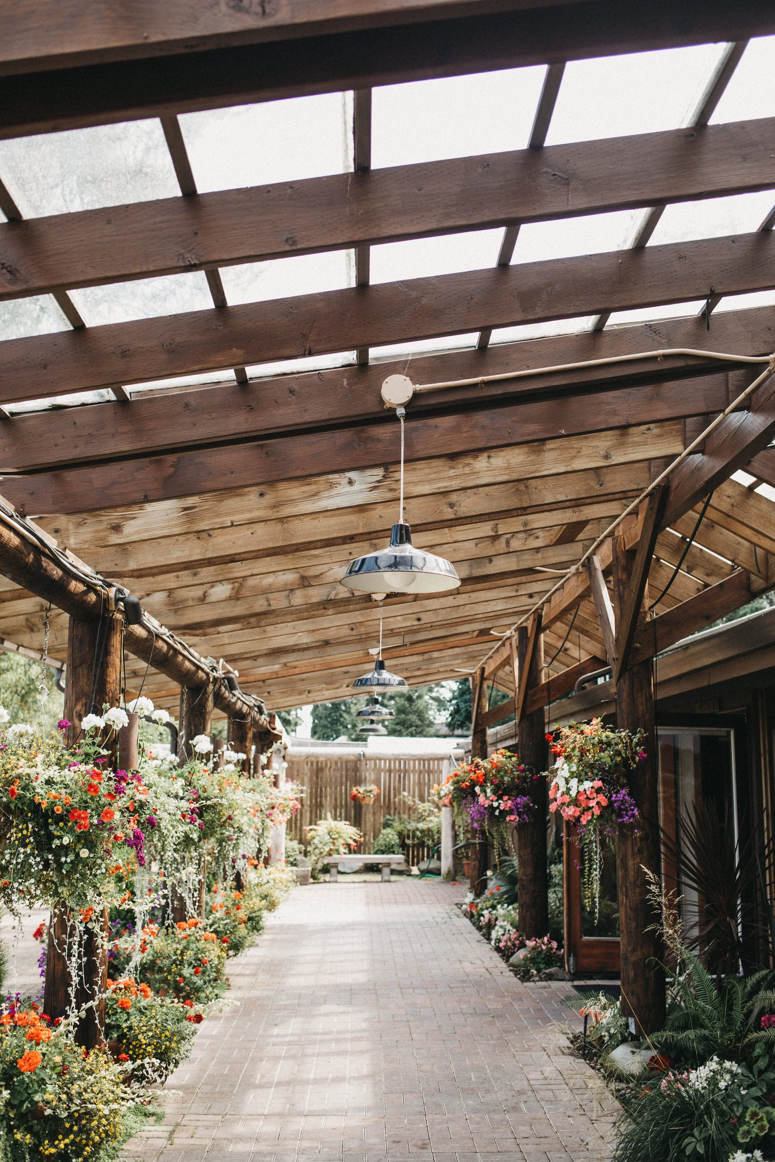 A covered outdoor walkway with hanging flower baskets and potted plants, wooden beams and a brick pathway.