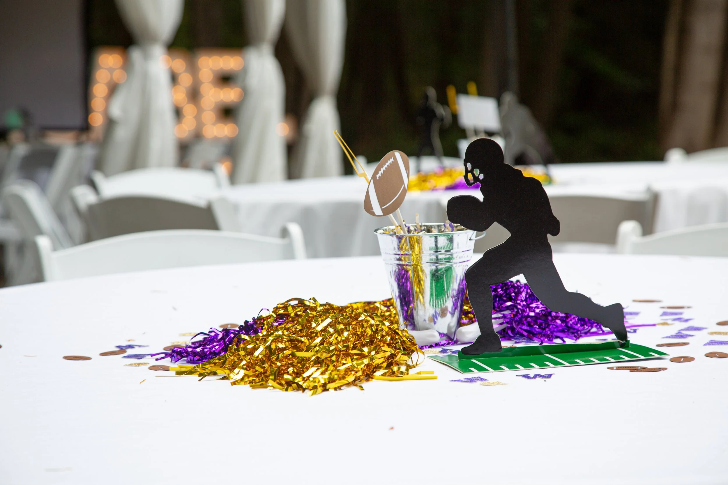 Table decoration with a football player silhouette, purple and gold confetti, and a metal cup with striped paper straws for a sports-themed celebration.