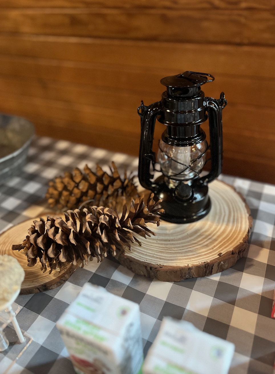 Decorative scene with a black lantern, large pinecones, and a wood slice on a checkered tablecloth.