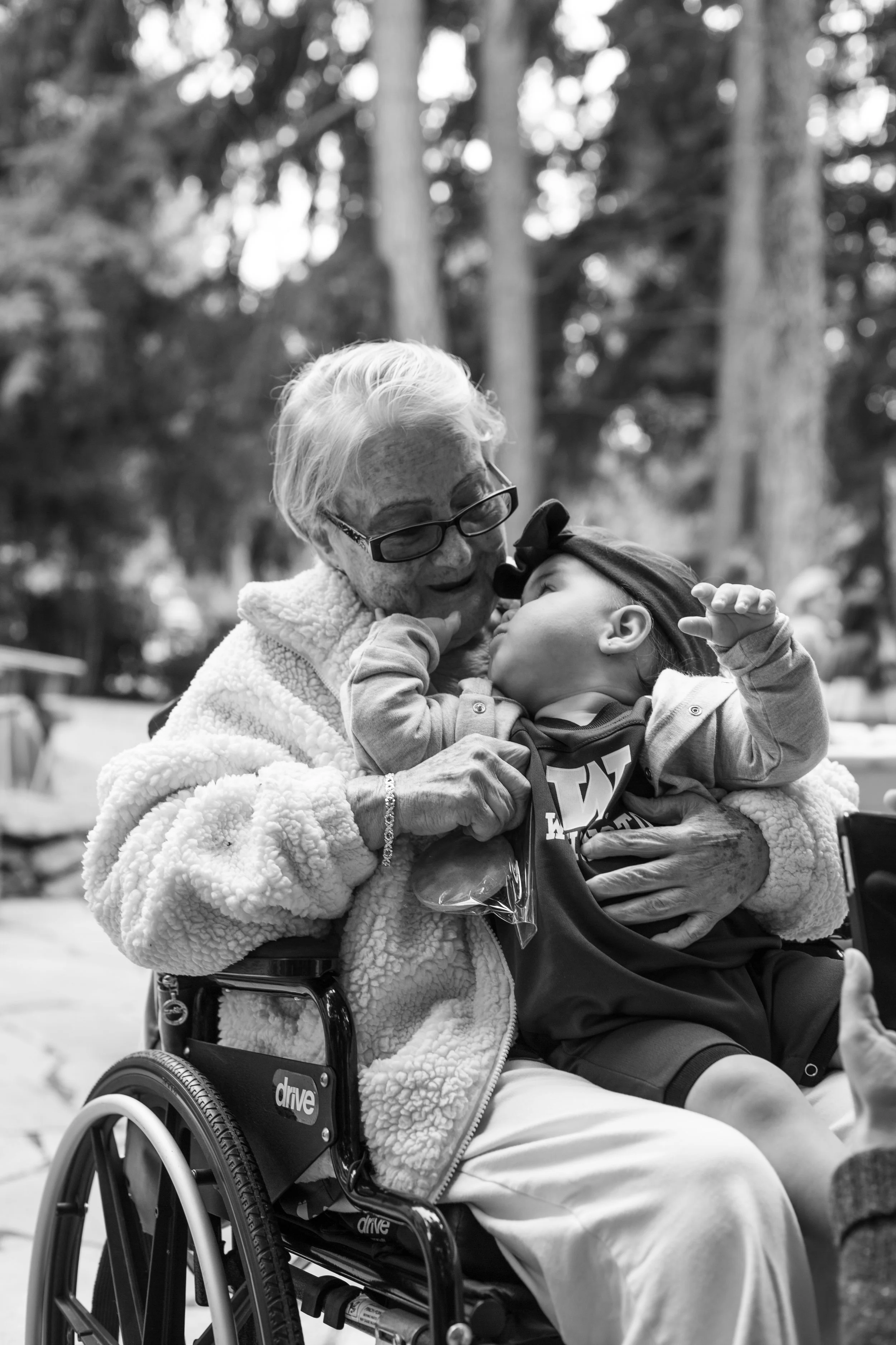 An elderly woman in a wheelchair holding a young child. The woman is smiling and wearing glasses, and the child is touching her face with his hand. They are outdoors with trees in the background.