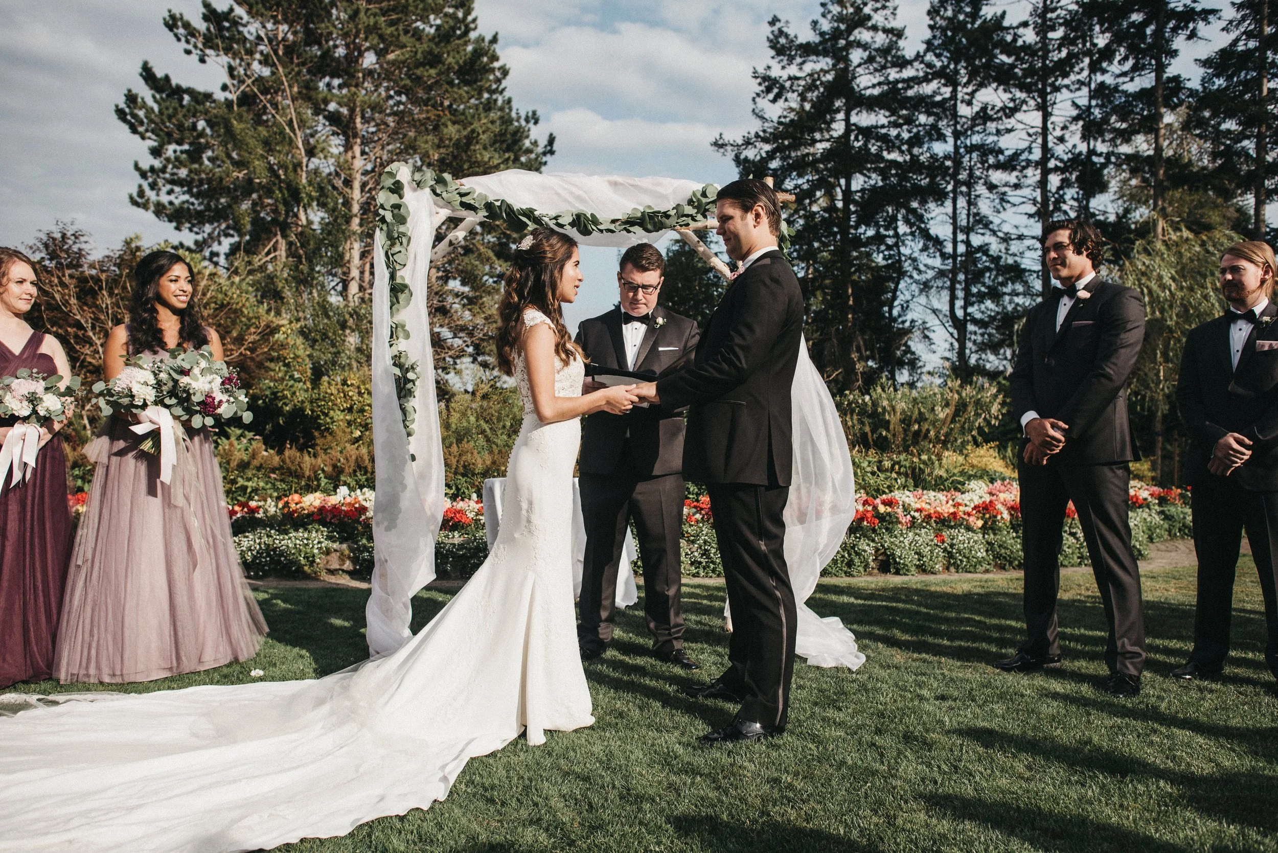 A couple getting married outdoors under a white floral arch while holding hands, with the officiant in the middle. Bridesmaids and groomsmen stand on either side, all dressed formally, in a garden with trees and colorful flowers.
