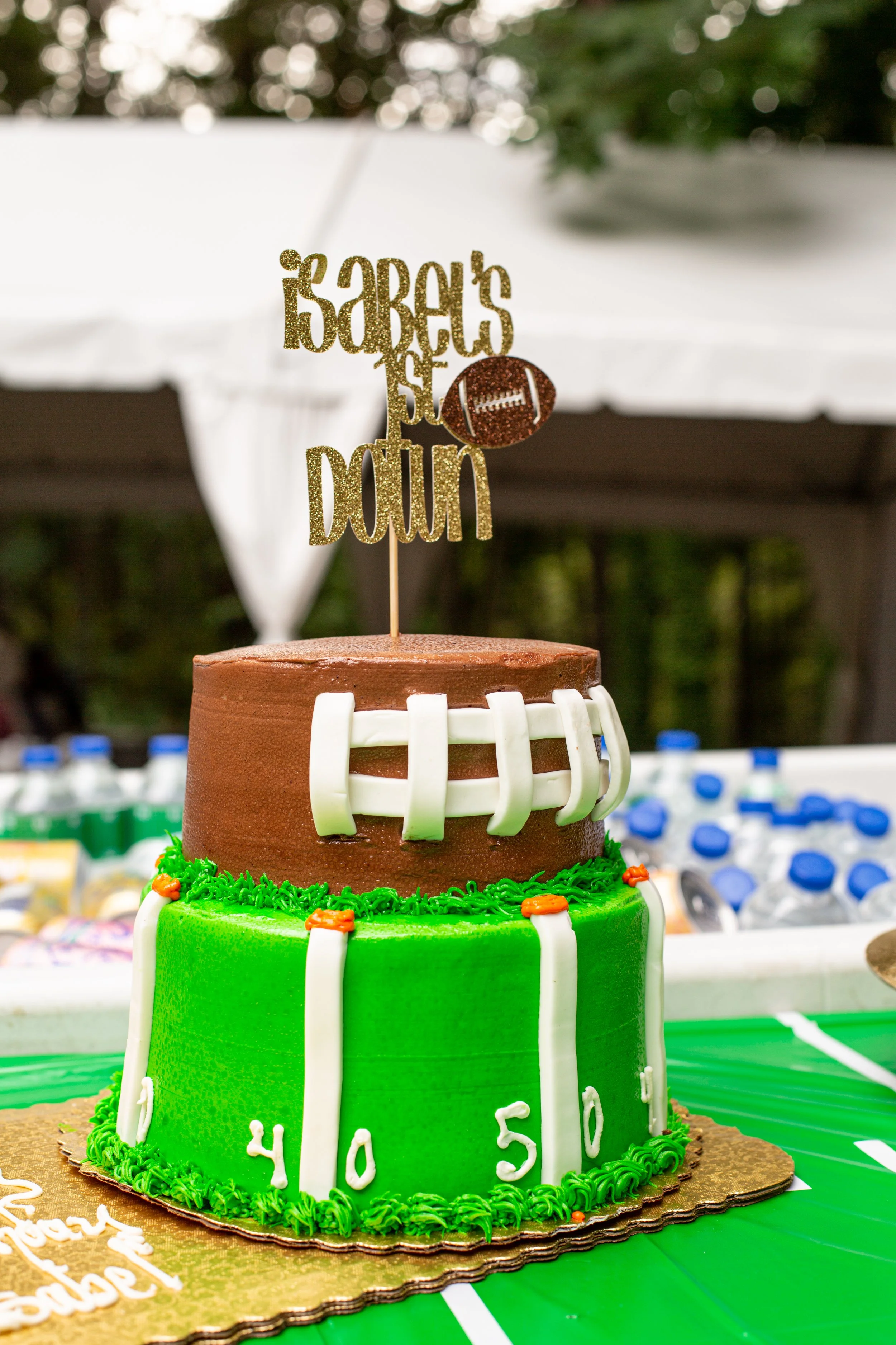Two-tiered football-themed birthday cake, with green base and brown top, decorated with white fondant to resemble a football and yard markers. The cake topper reads 'Isaiah's 5th Down' with a football graphic, set on a green tablecloth, with water bo