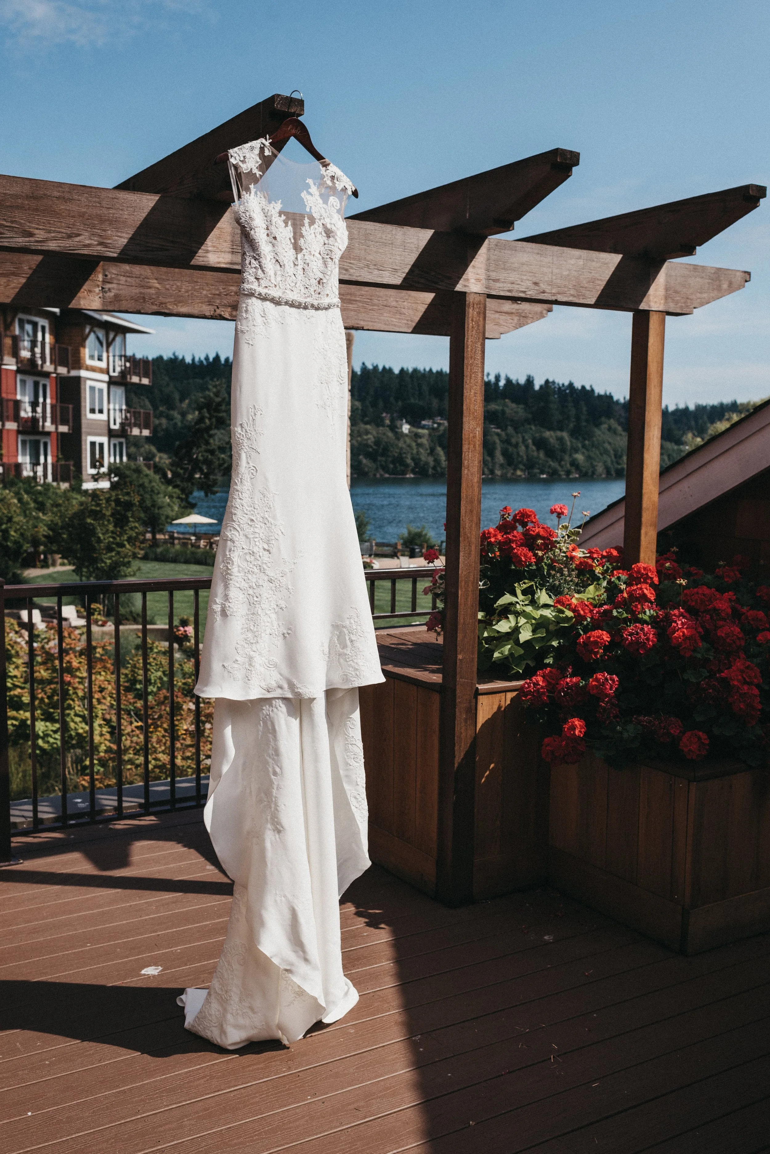 A white wedding dress hanging on a wooden beam on an outdoor balcony with a view of a lake, trees, and red flowers.