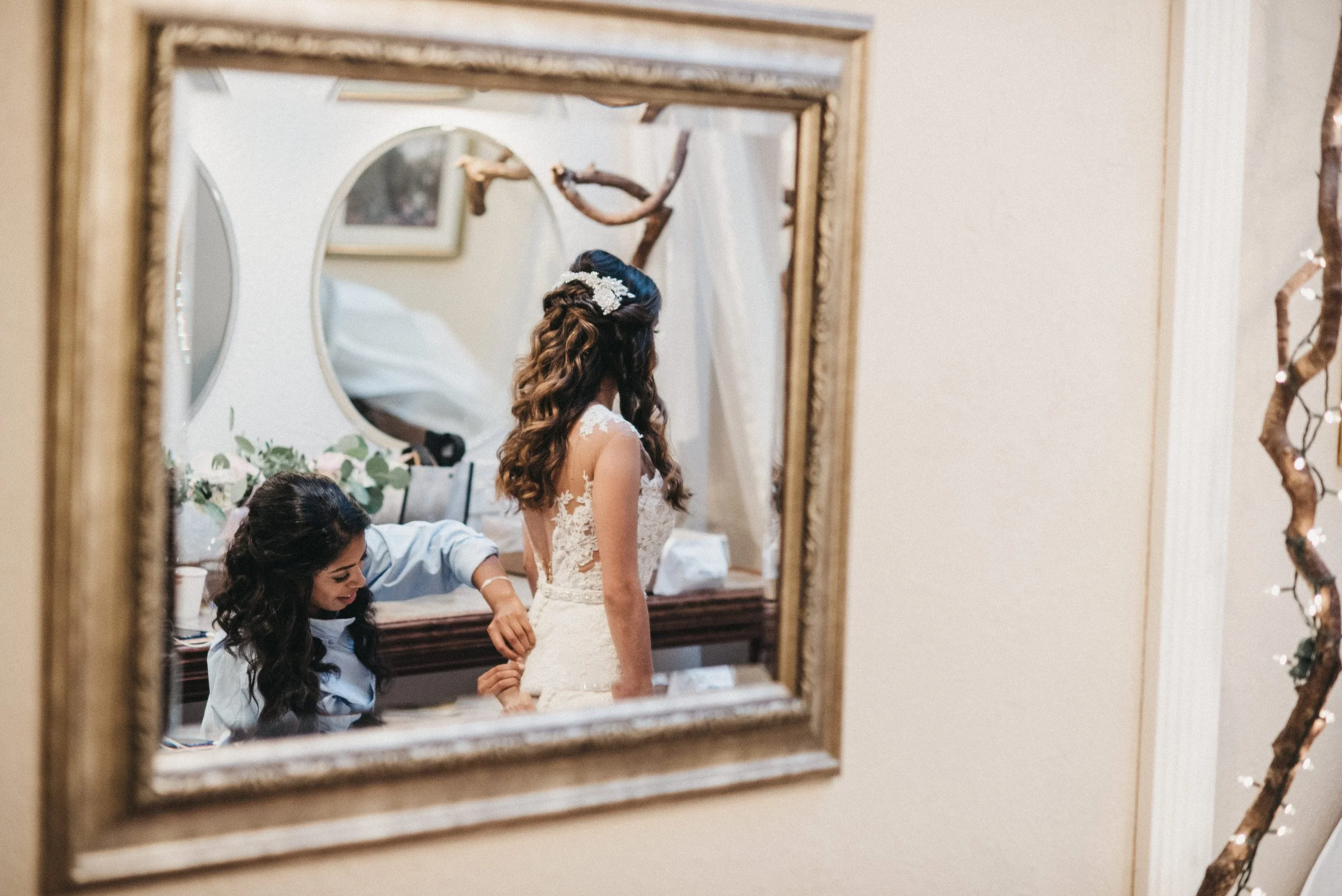 Two women preparing a bride for her wedding, one adjusting her dress, reflected in a mirror with a decorative frame, with natural and soft lighting.
