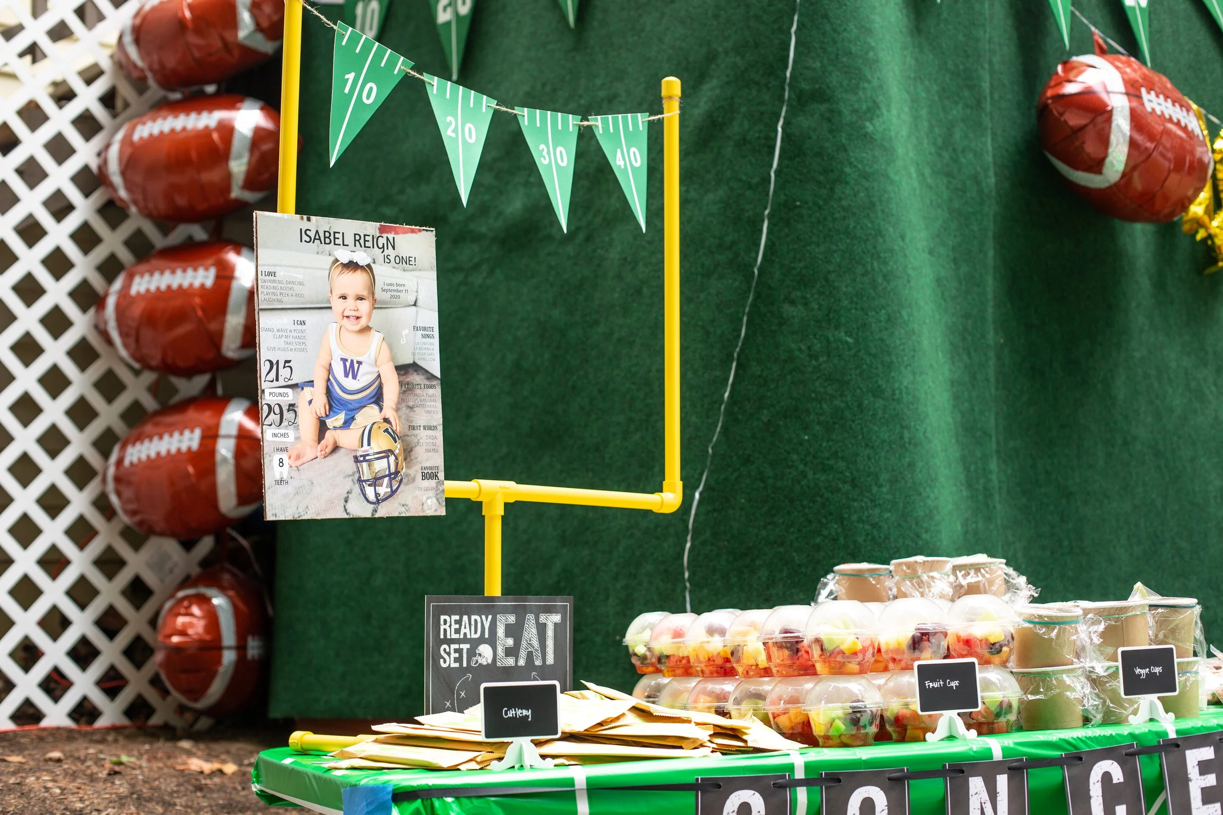 A table at a birthday celebration with bowls of fruit cups, veggie cups, and chopped fruit, with a sign saying 'Ready, Set, Eat.' In the background, there's a green wall with football-themed decorations, including red foil football balloons, a hangin