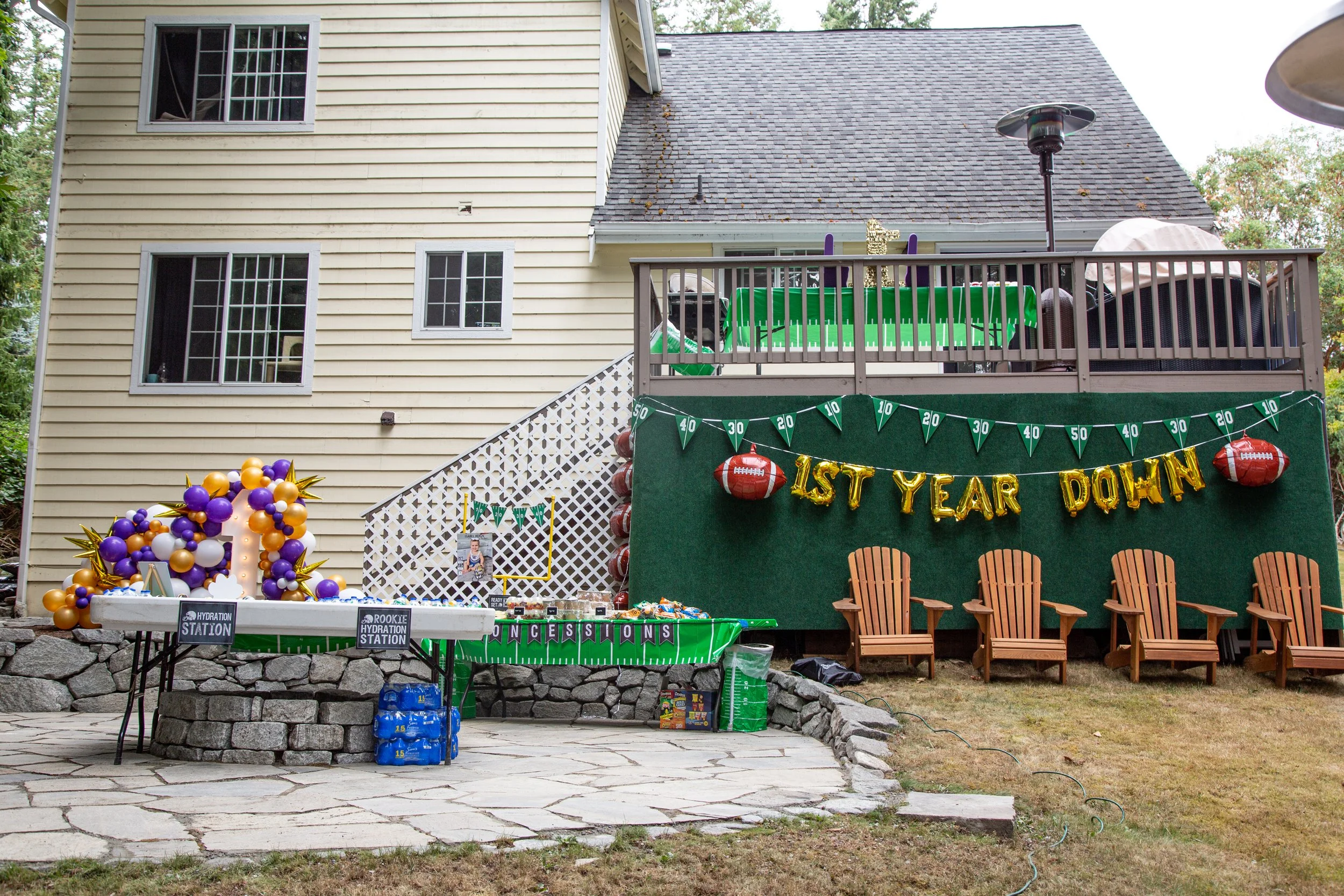 Backyard party setup with a decorated green banner that reads '1st Year Down', football-themed balloons, and a table labeled 'hydration station' with purple, gold, and white balloons shaped in the number 1. There are four wooden chairs on the grass i