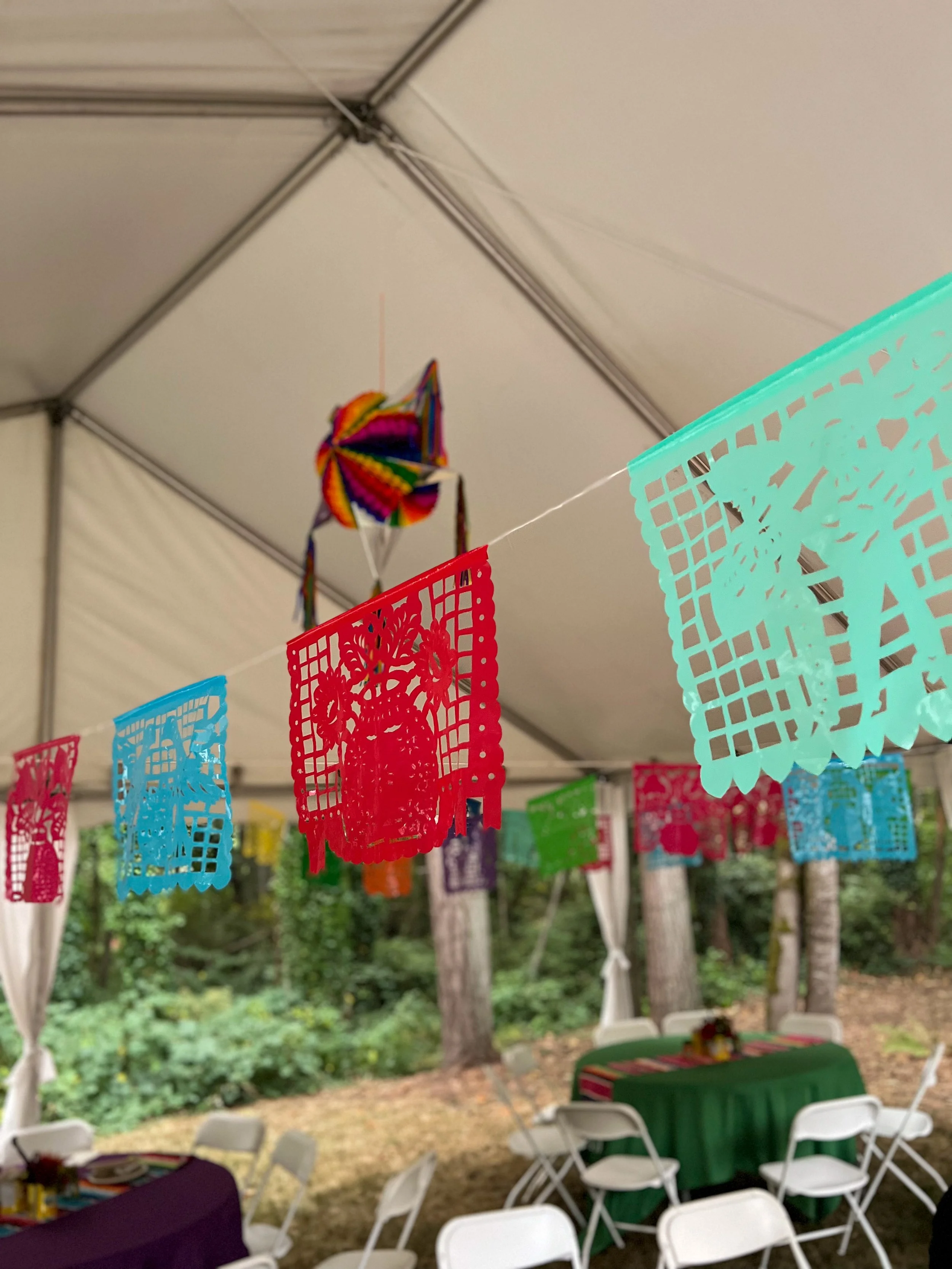 Colorful papel picado banner hanging under a white party tent. Inside the tent, there are tables with green and purple tablecloths and white chairs, set for a celebration in an outdoor wooded area.