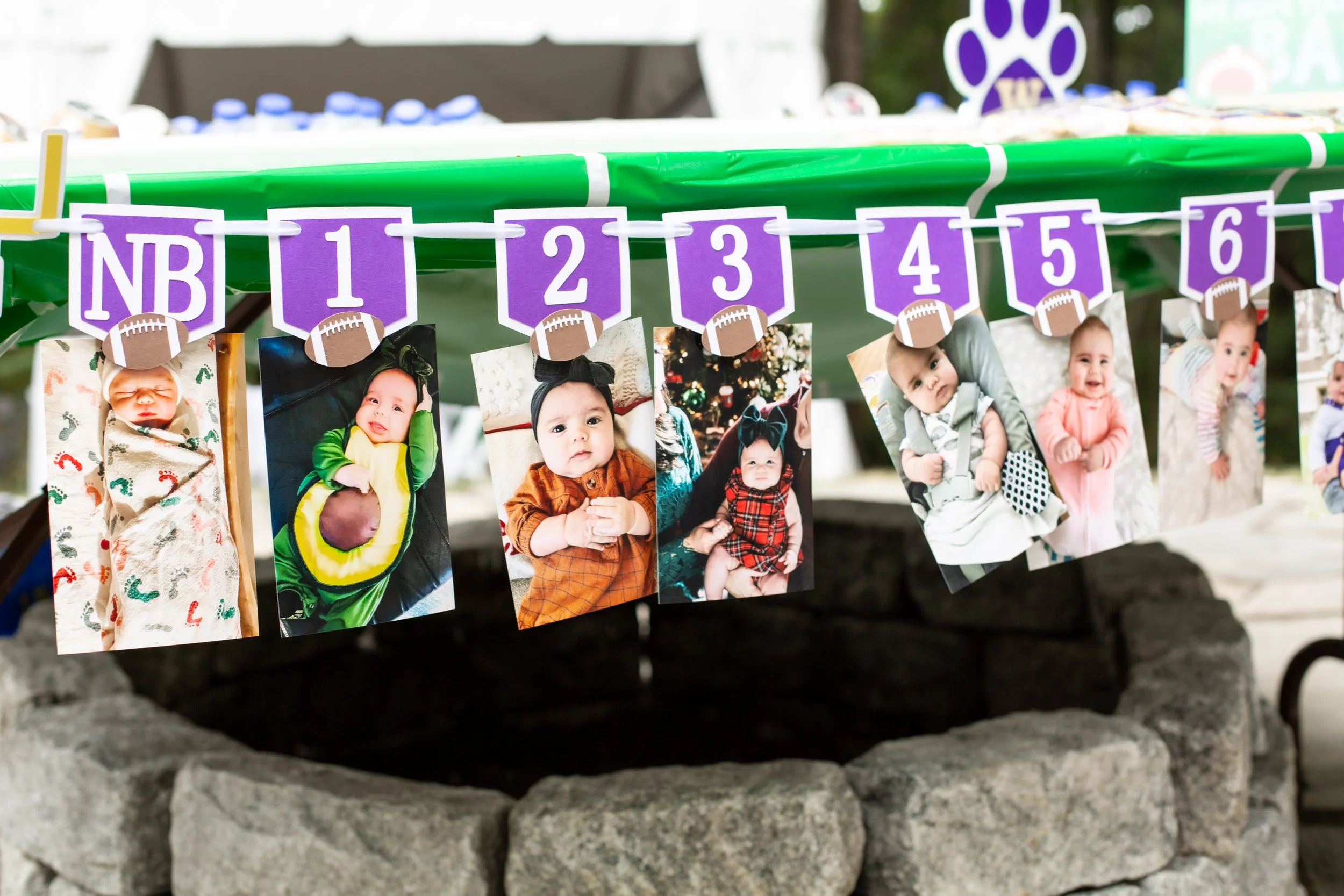 A display of seven printed photos of a baby girl, hanging on a string with purple and white signs above, numbered NB to 6, with football-themed decorations, in front of a stone circle. The photos show the baby in different outfits and poses.