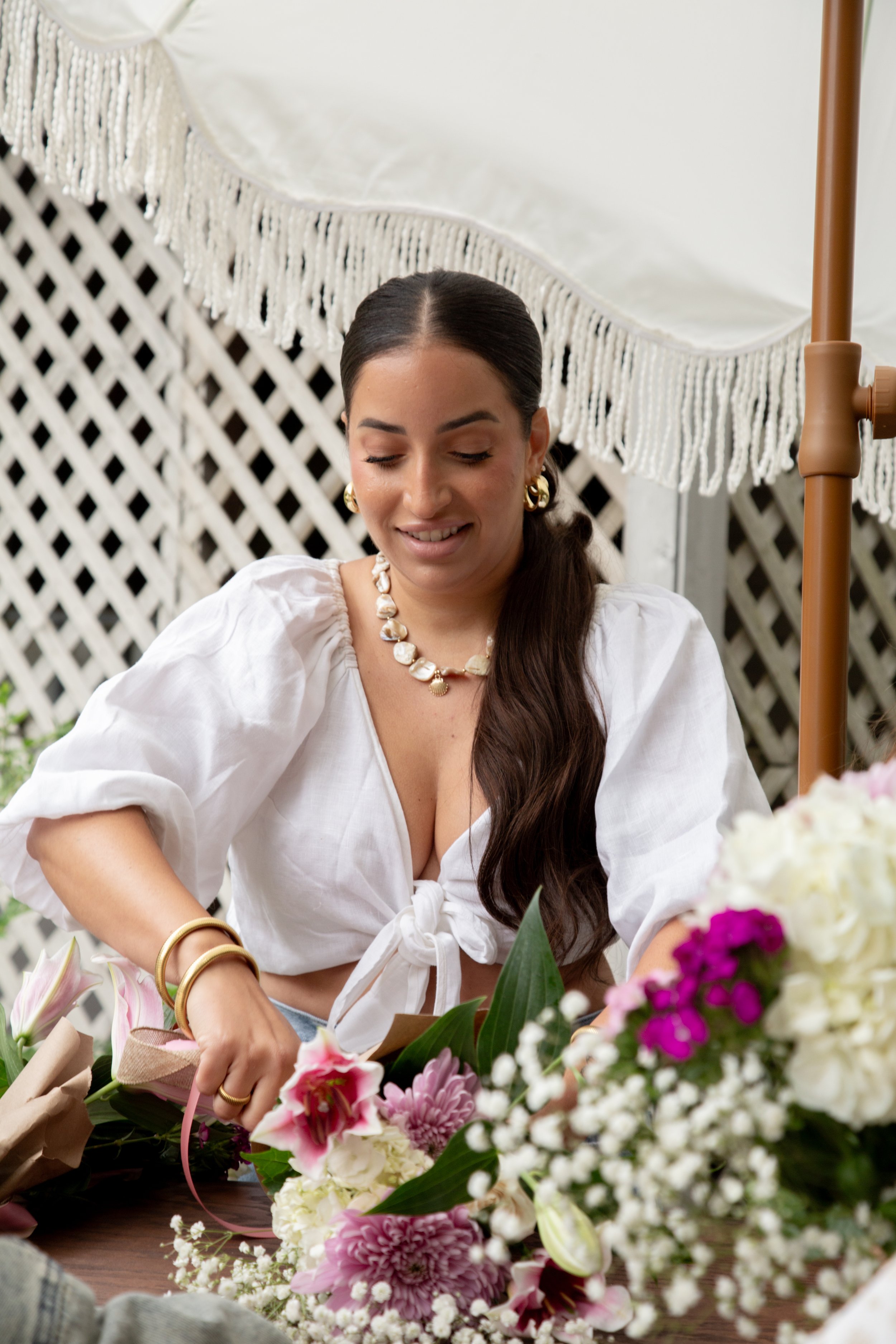 A woman with dark brown hair wearing gold jewelry, a white blouse, and gold bangles, smiling with her eyes closed while arranging flowers at a table with a variety of pink, white, and purple flowers, under a white umbrella with fringe.