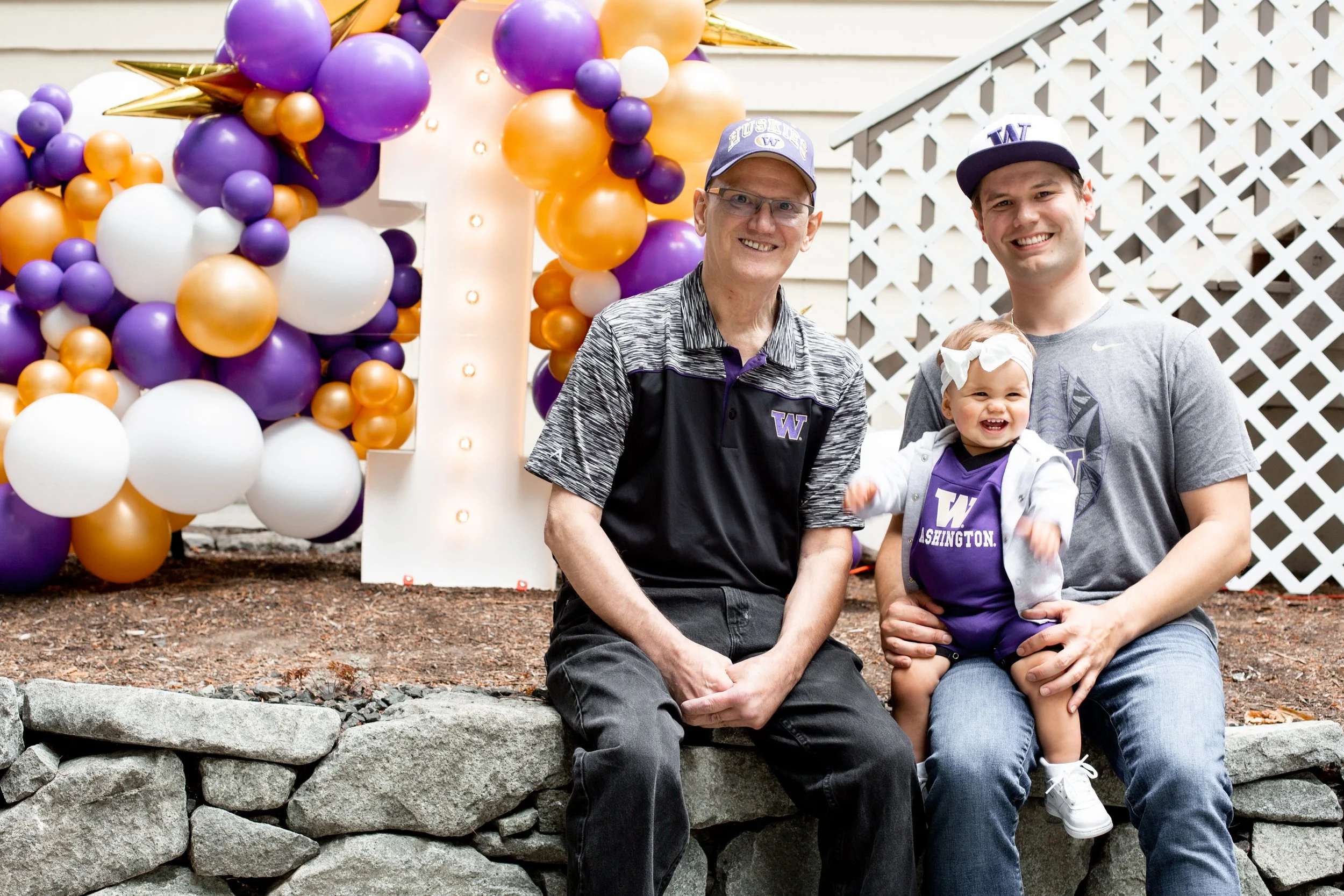 A family of three celebrating outdoors with purple, white, and gold balloons and a large illuminated number one. The grandfather, father, and young girl are sitting on a stone wall, smiling at the camera. The girl is wearing a Washington Huskies outf