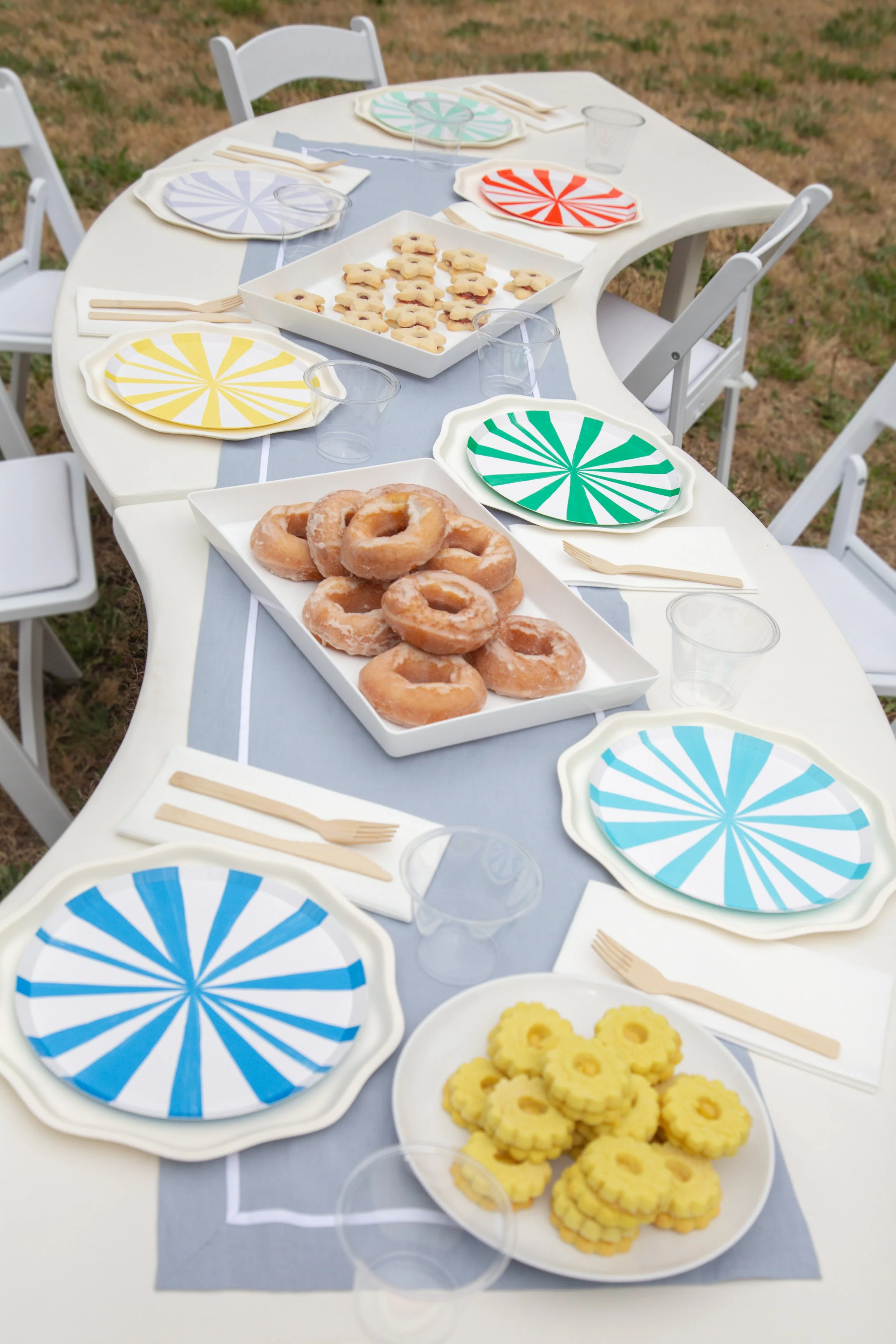 A table set for a celebration with colorful paper plates, some with a radiating stripe pattern in yellow, blue, green, and red, accompanied by wooden cutlery, clear plastic cups, and a gray table runner. The table features plates of donuts, sugar cookies, and candies, indicating a festive or casual gathering outdoors with grass visible in the background.