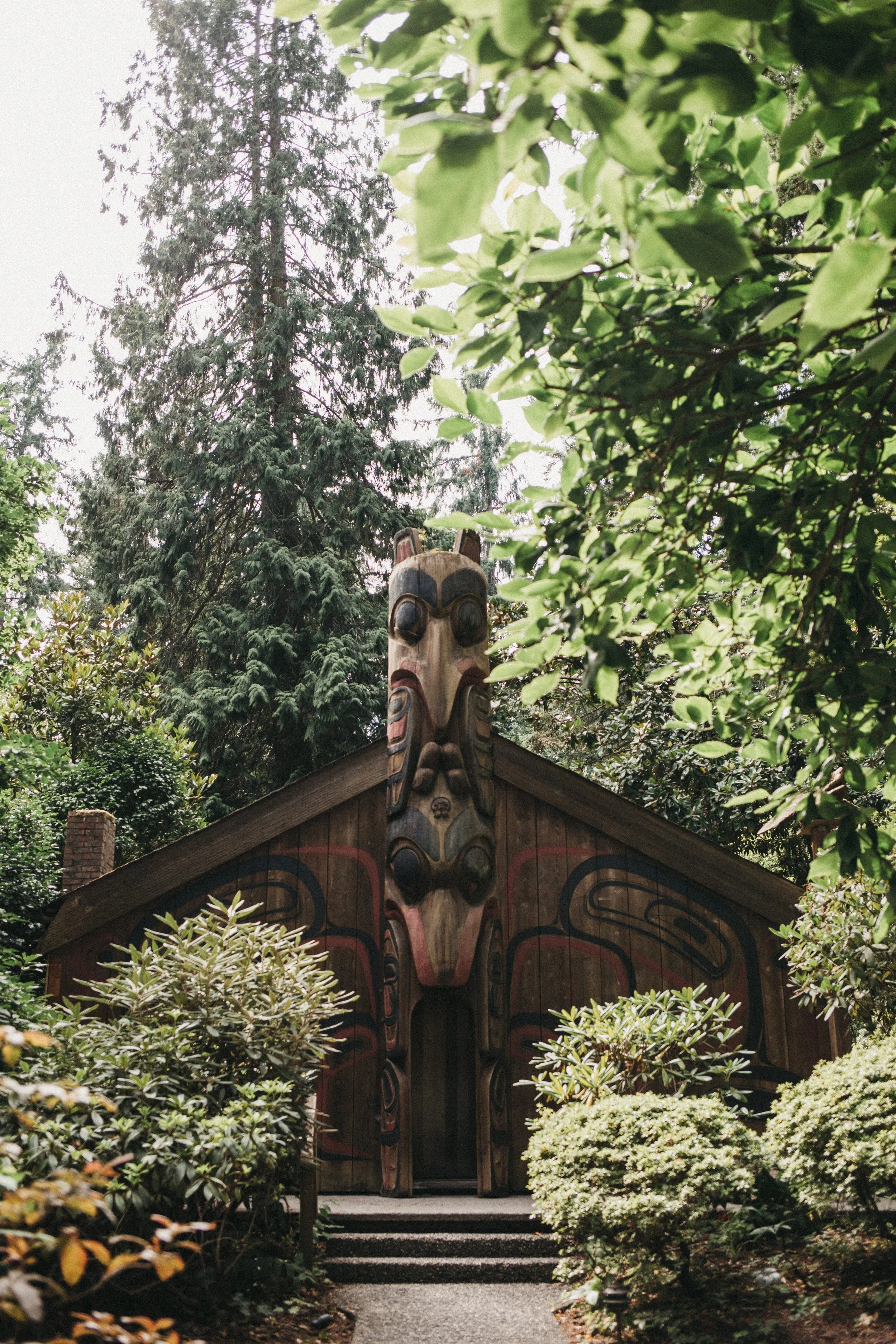 A traditional indigenous house with a large carved wooden totem pole on its roof, surrounded by lush green trees and bushes.