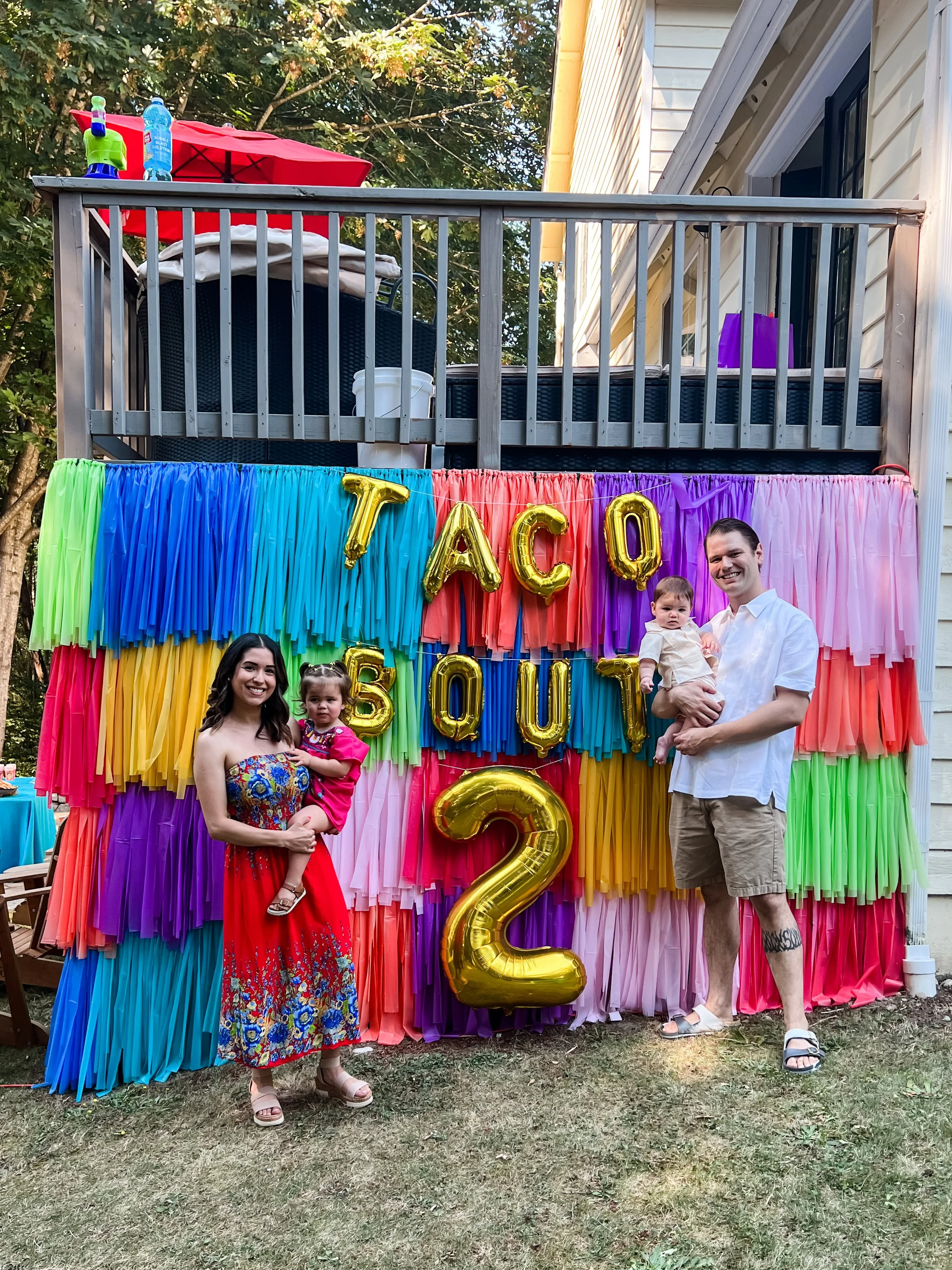 Family celebrating a second birthday in backyard with colorful paper backdrop, balloon letters spelling 'TACO BOUT 2', and two adults each holding a child.