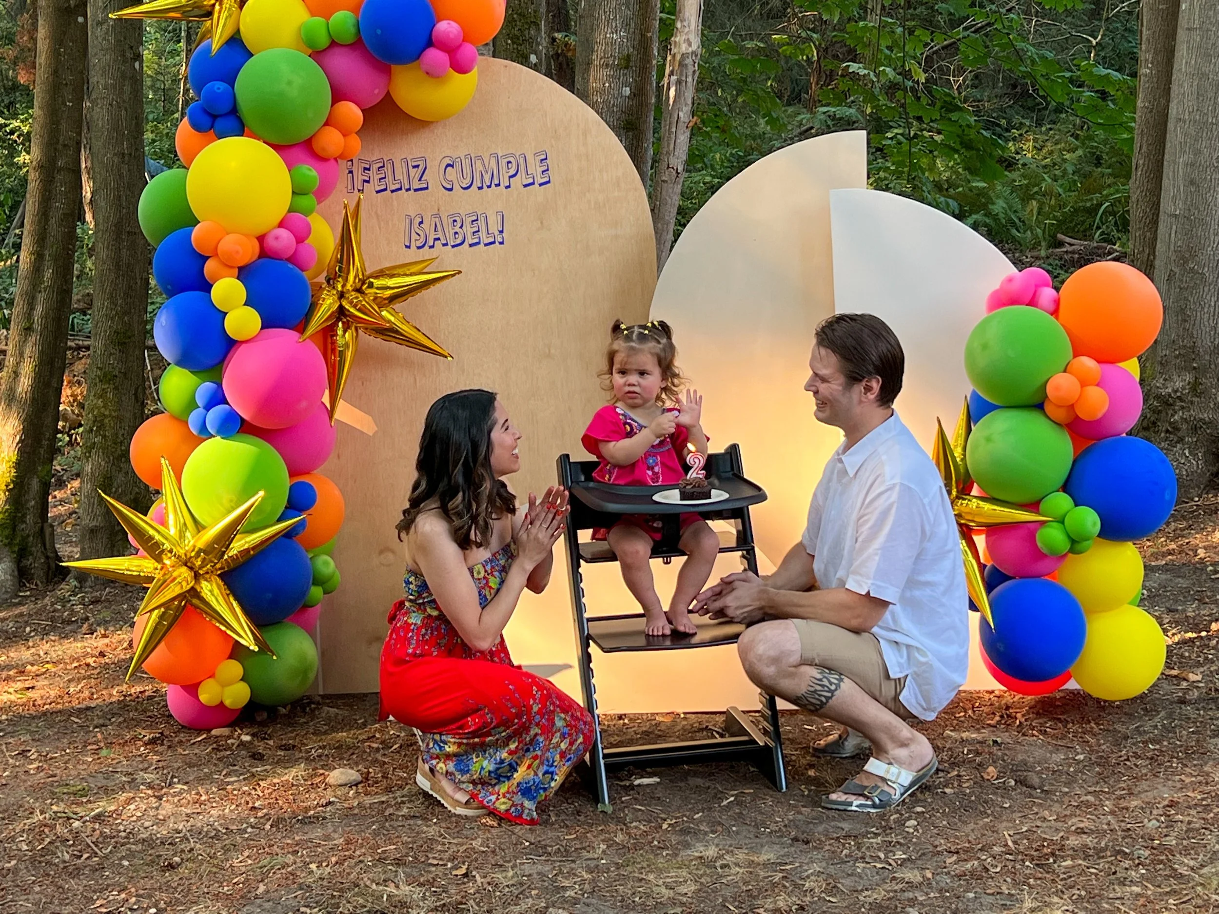 A young girl at her birthday celebration outdoors in a wooded area, sitting in a high chair with a cake, past a backdrop with colorful balloon decorations and a birthday message in Spanish, flanked by two adults.