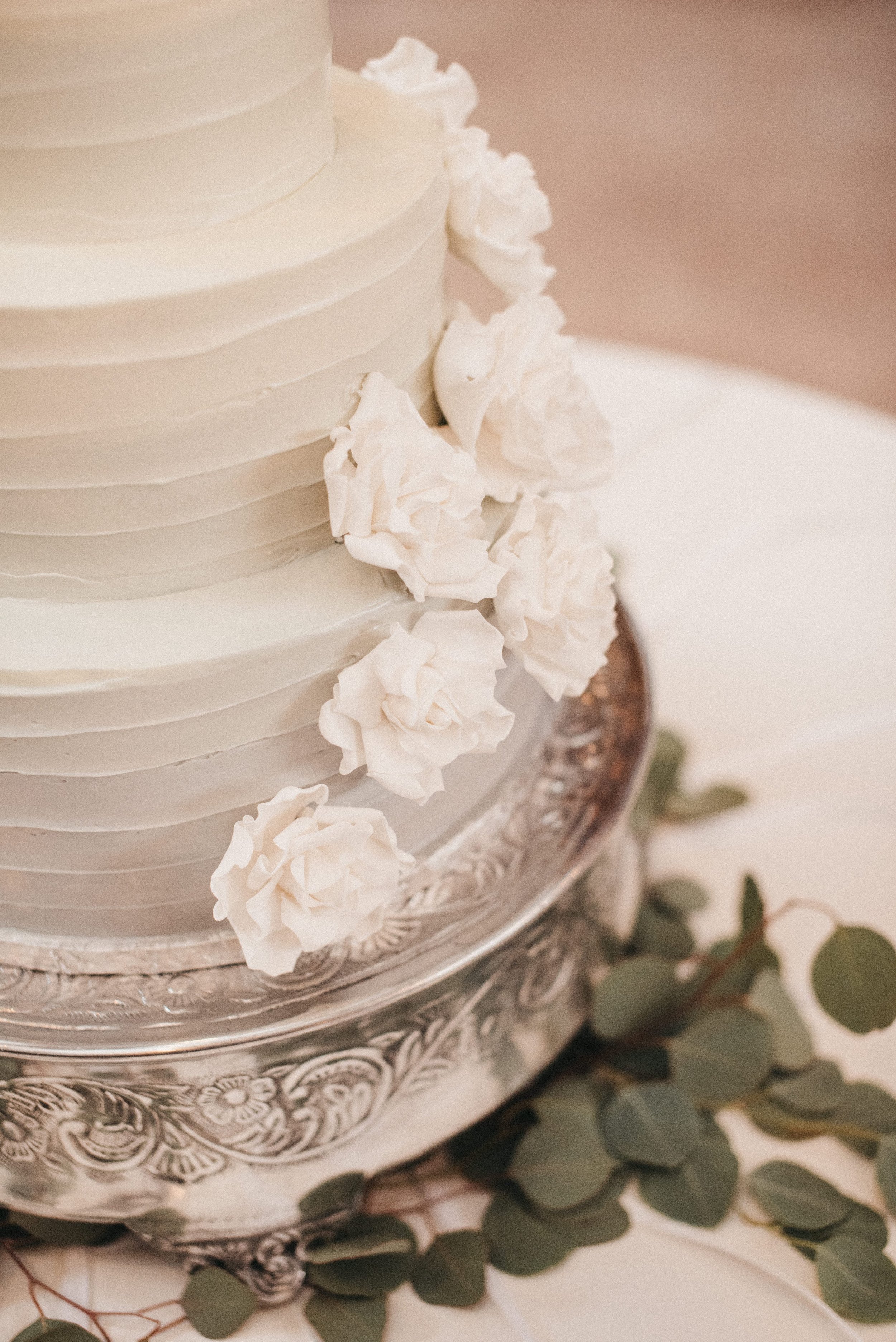 A tiered wedding cake decorated with white flowers, placed on a silver ornate cake stand with eucalyptus leaves surrounding it.