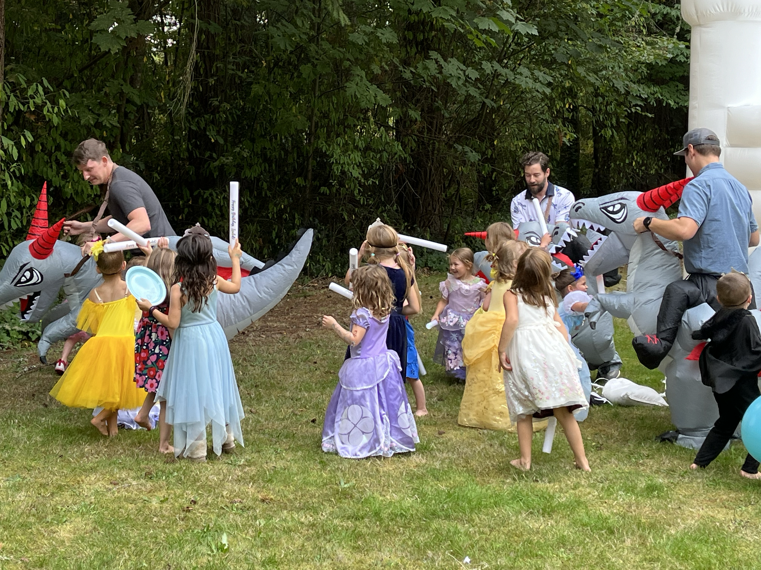 Children and adults at a party outdoors, playing with inflatable dragon toys, dressed in colorful party attire, on a grassy area next to a wooded background.