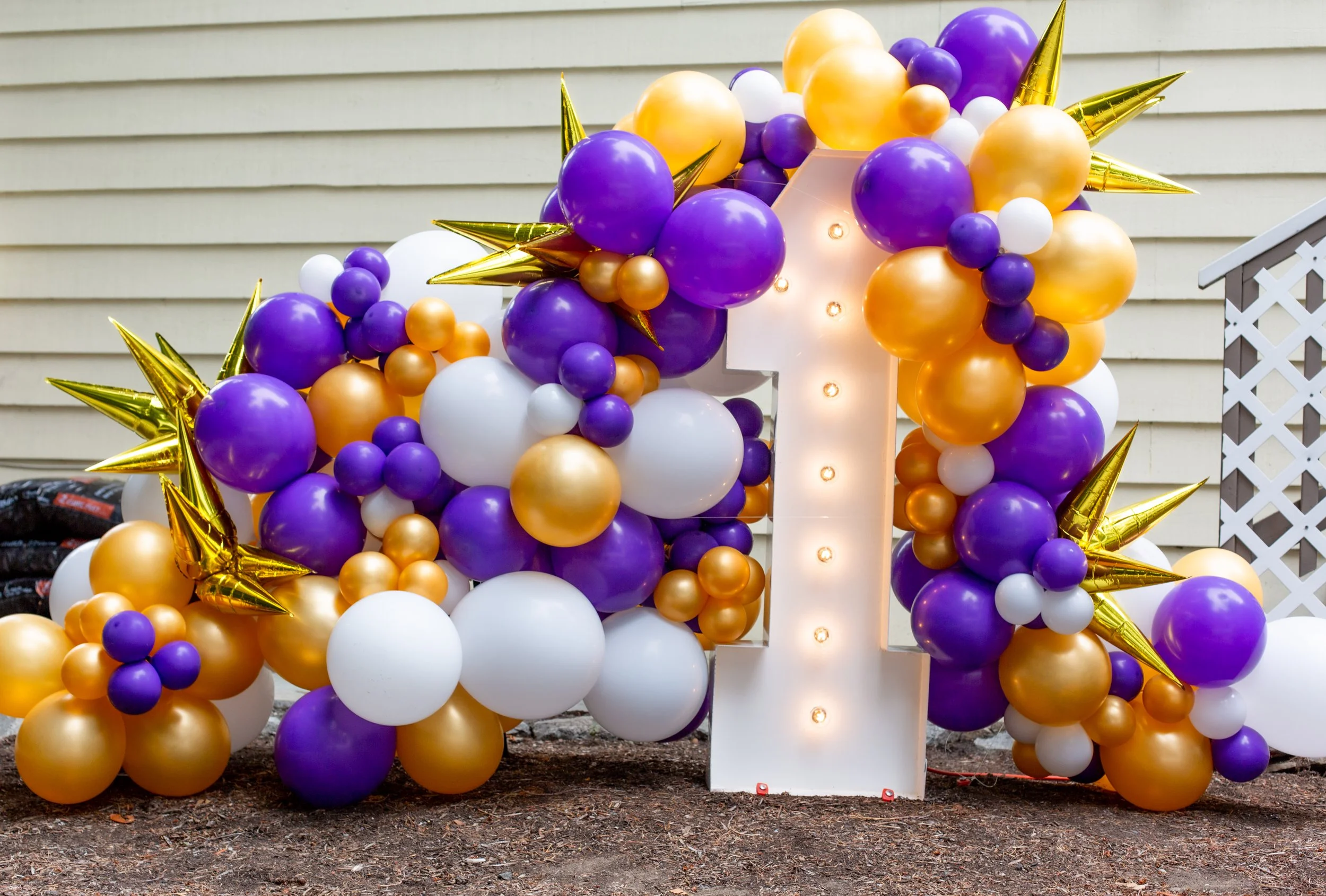 Decorative balloon display with purple, white, and gold balloons densely arranged around a large illuminated number one on a stand.