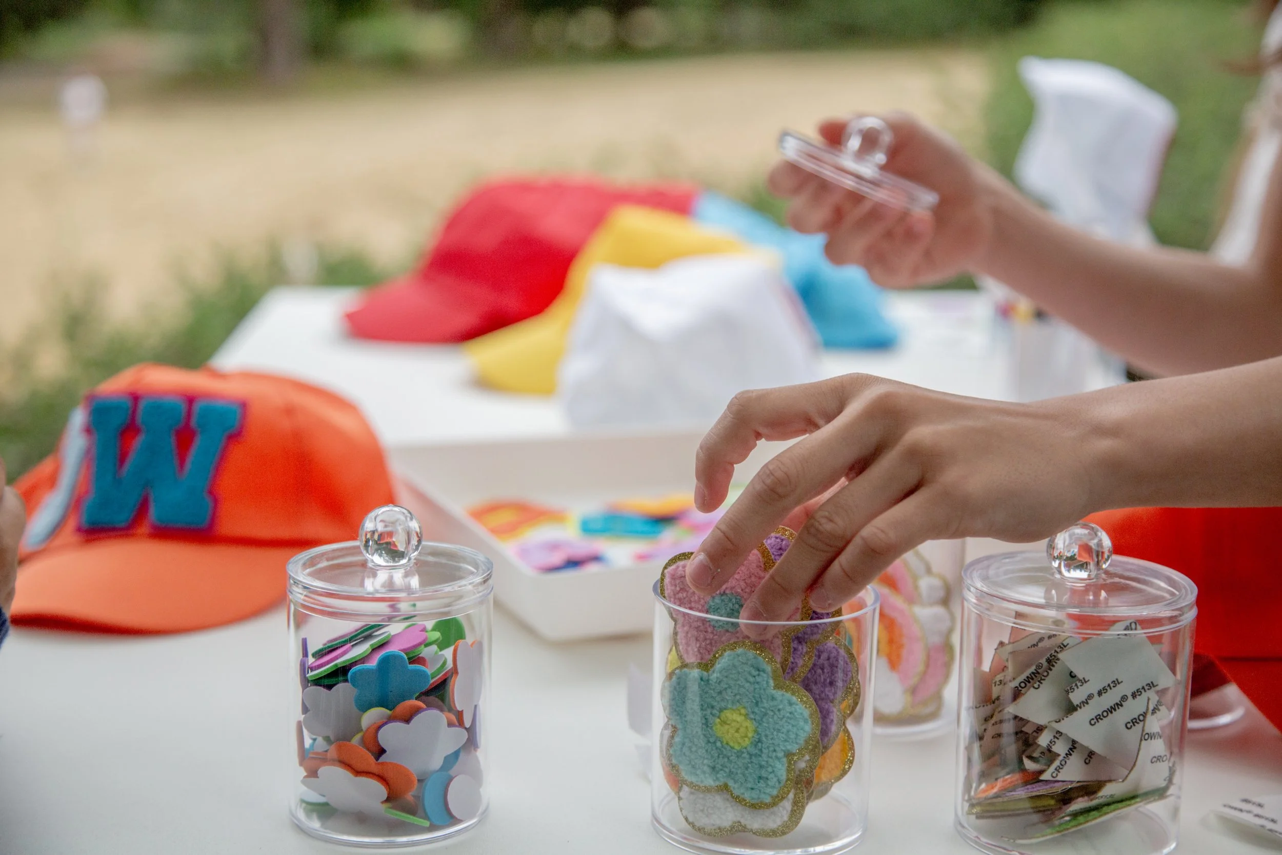 Close-up of hands reaching into glass jars filled with colorful craft supplies, including foam shapes, glittery cutouts, and small decorated cookies, with a blurred outdoor park in the background.