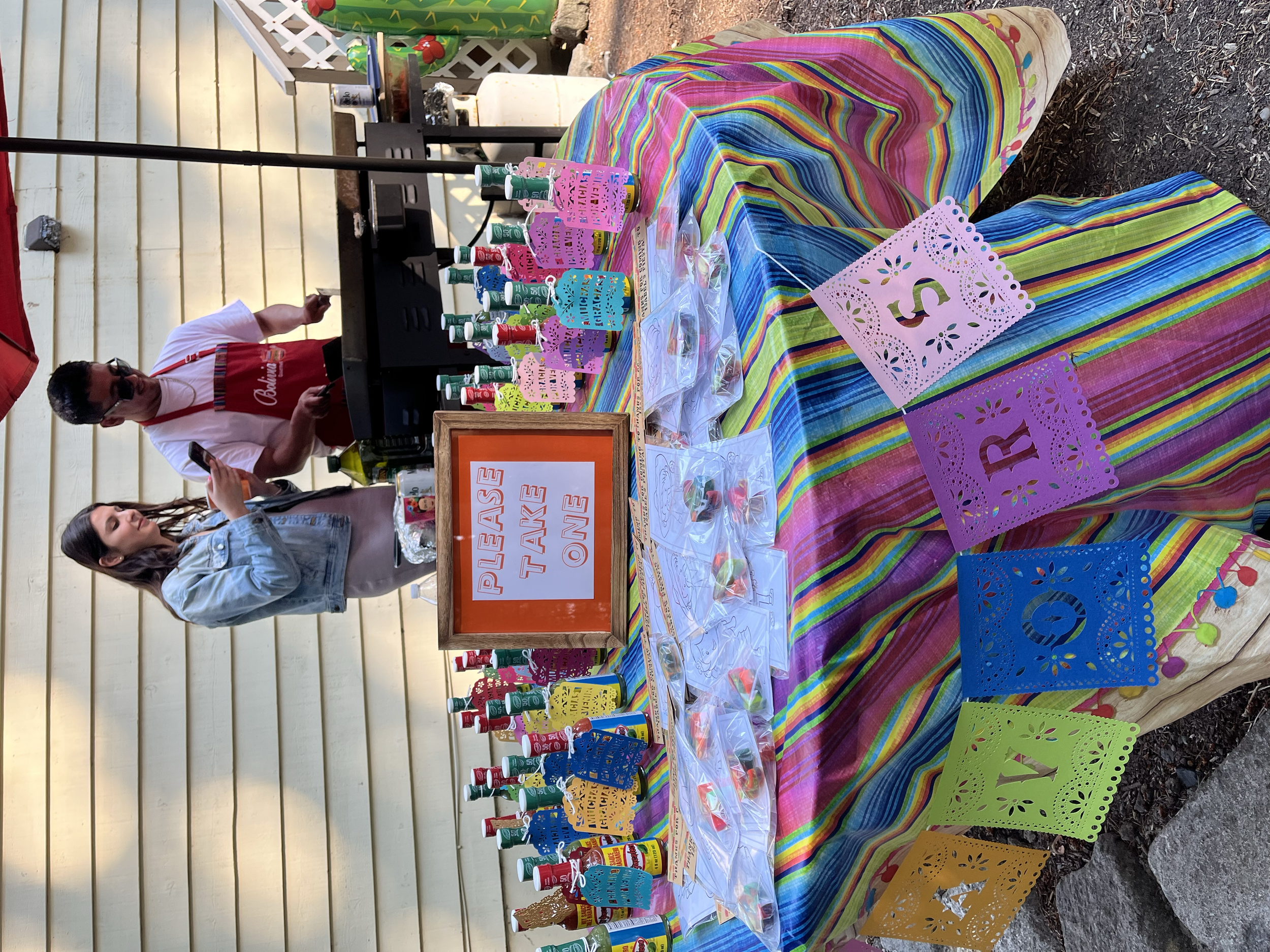 Colorful table with bottles of hot sauce, paper banners with letters spelling 'SUR' and 'FIR', and a 'Please Take One' sign. Two people are behind the table, one wearing sunglasses and a red apron, the other taking a photo. The table is covered with 