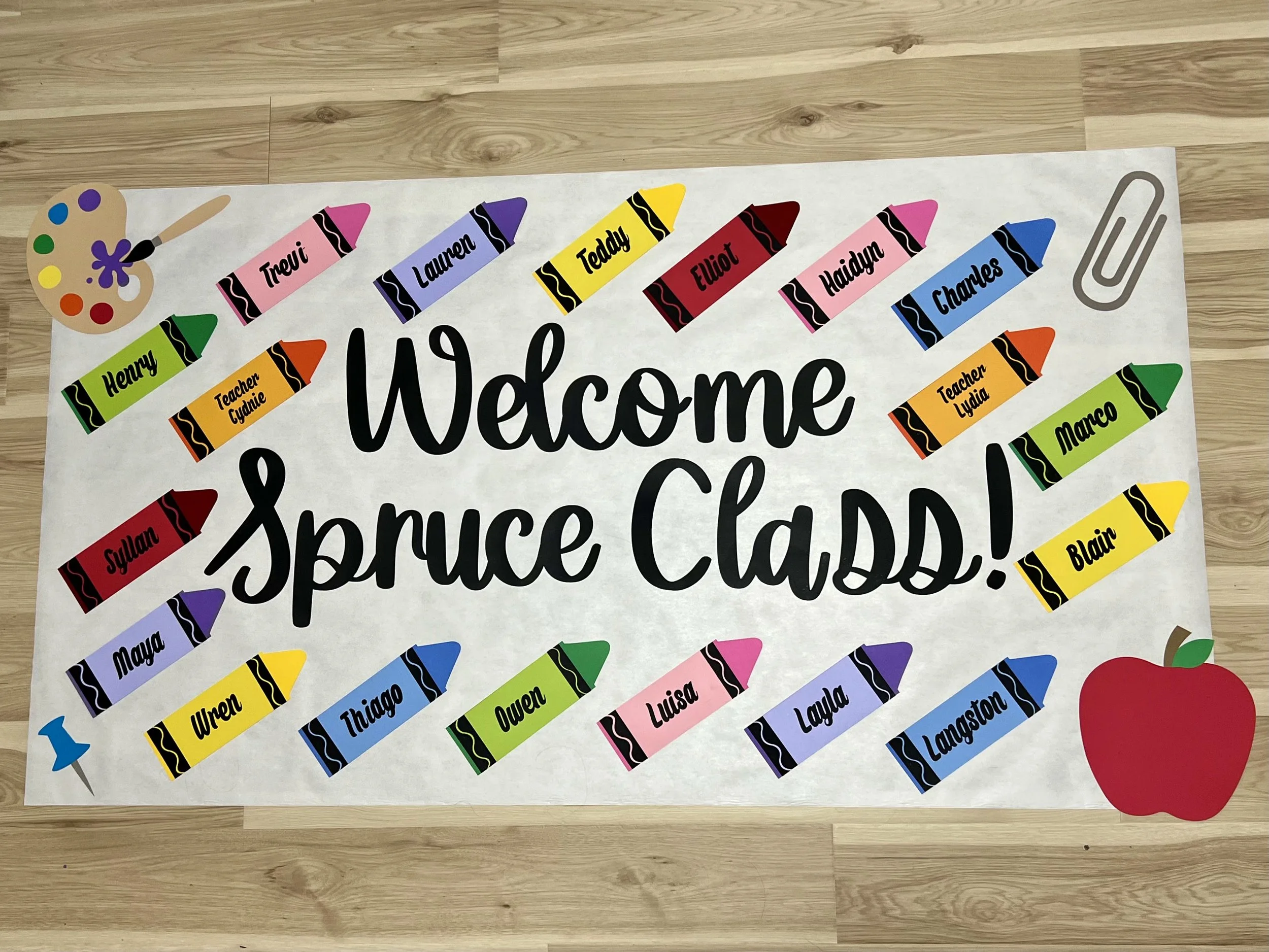Decorative classroom poster with the message 'Welcome Spruce Class!' surrounded by paper crayon cutouts labeled with students and teachers' names, a paperclip, and a small artist's palette with a paintbrush.