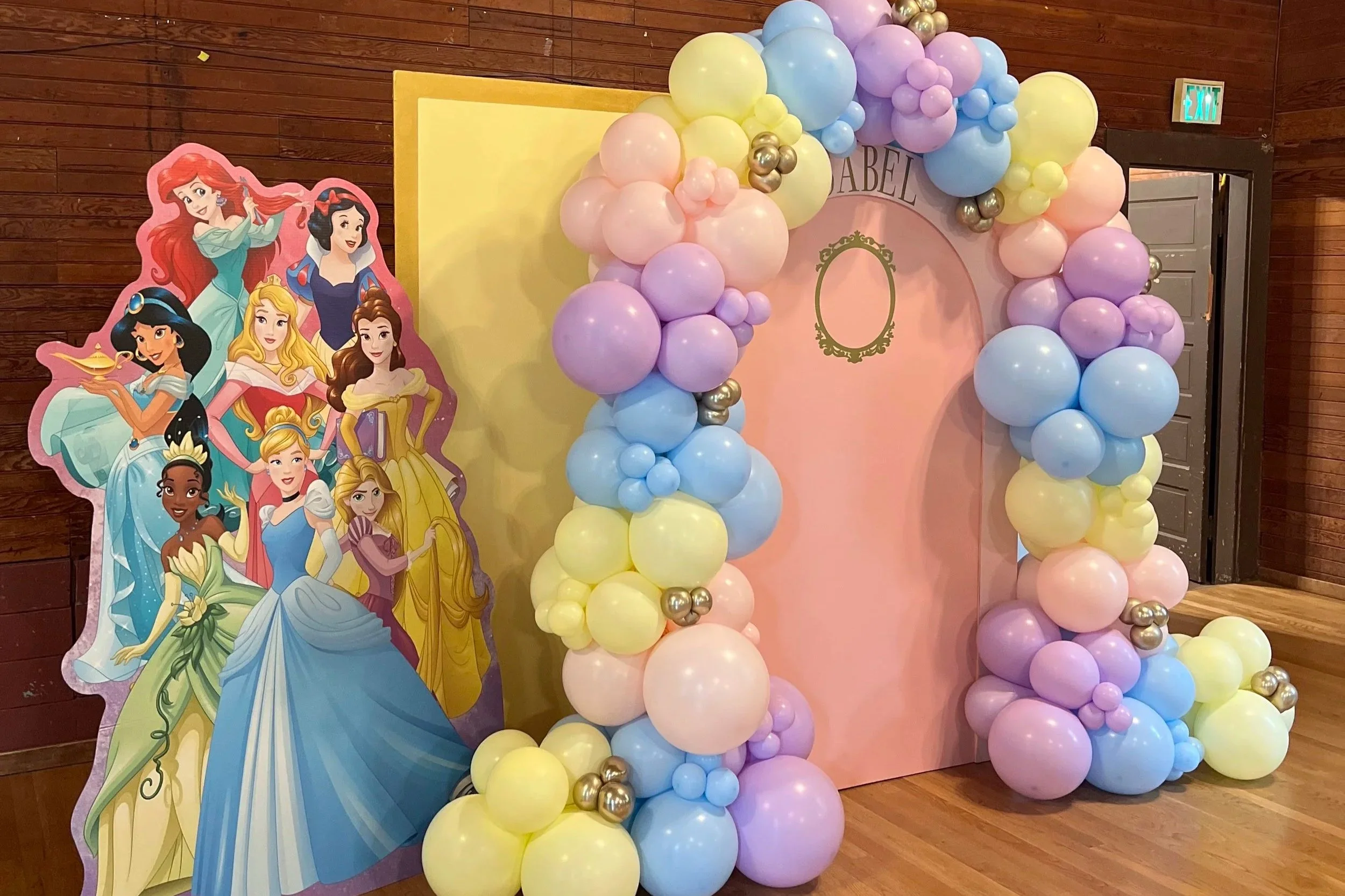 Colorful balloon arch with pastel pink, blue, yellow, purple, and silver balloons surrounding a pink backdrop with a gold frame outline. To the left, a cardboard cutout display features Disney princess characters including Ariel, Snow White, Belle, Cinderella, and others.
