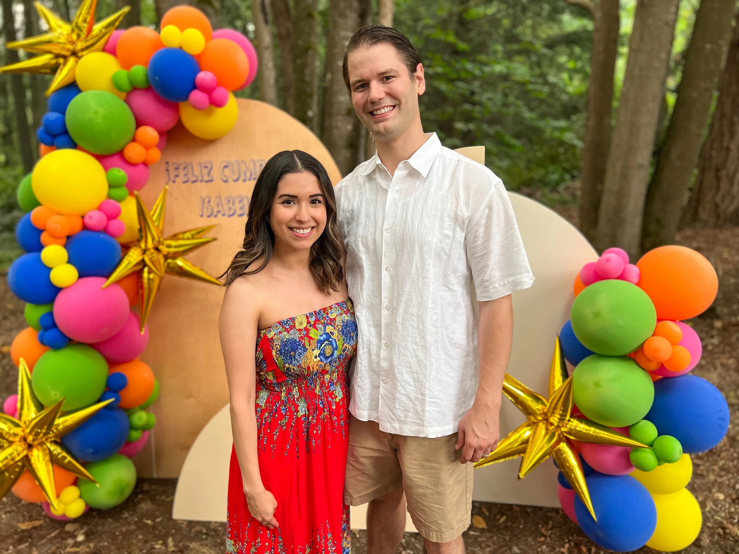 A smiling woman in a strapless colorful floral dress and a tall man in a white button-up shirt and khaki shorts stand together outdoors in front of a colorful balloon arch with stars, with trees in the background.