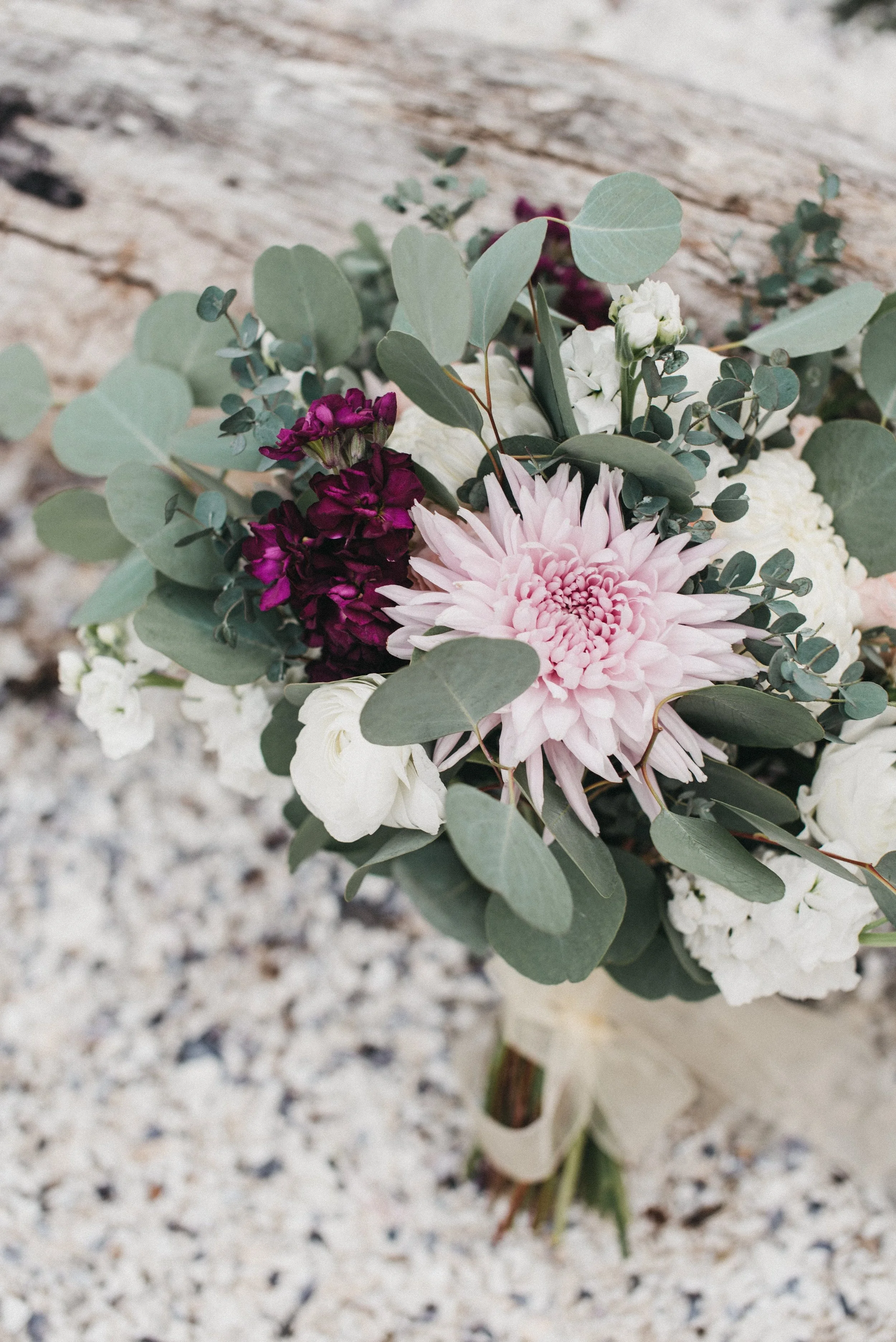 Close-up of a wedding bouquet with pink, purple, white flowers and green eucalyptus leaves, resting on white gravel with a weathered wooden surface in the background.