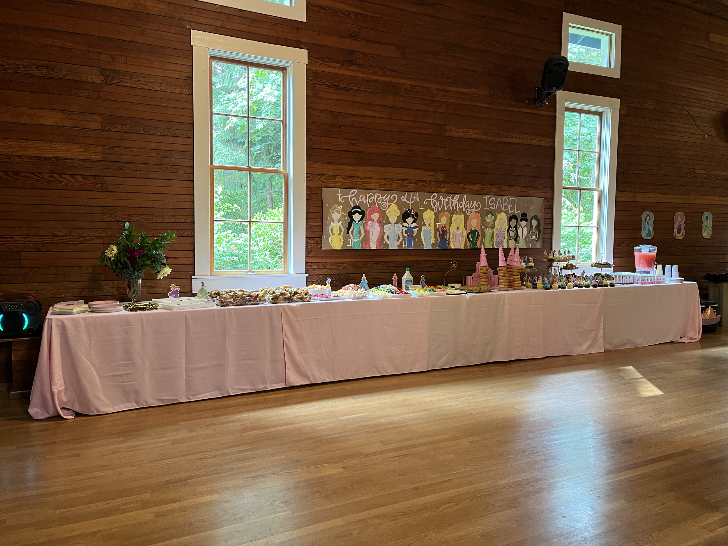 Long buffet table with pink tablecloth featuring birthday cake, snacks, pink cone-shaped party hats, desserts, punch drink, and party supplies in a rustic room with wooden walls and large windows.