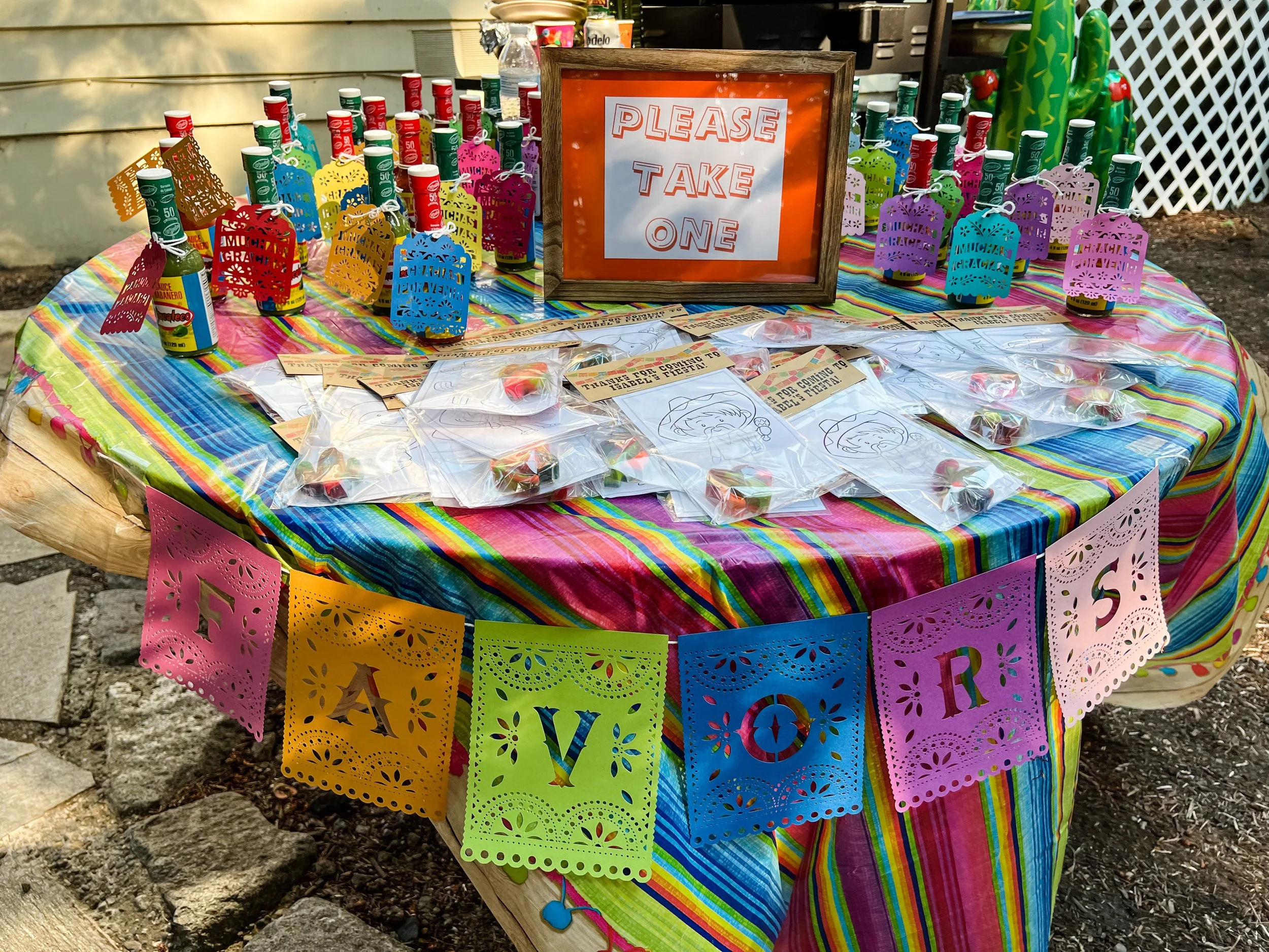Colorful fiesta table decorated with a rainbow striped tablecloth, featuring bottles with festive tags, a framed sign that says 'Please Take One,' and small bags with candies. A vibrant paper banner spelling 'FAVOR' decorates the front of the table.