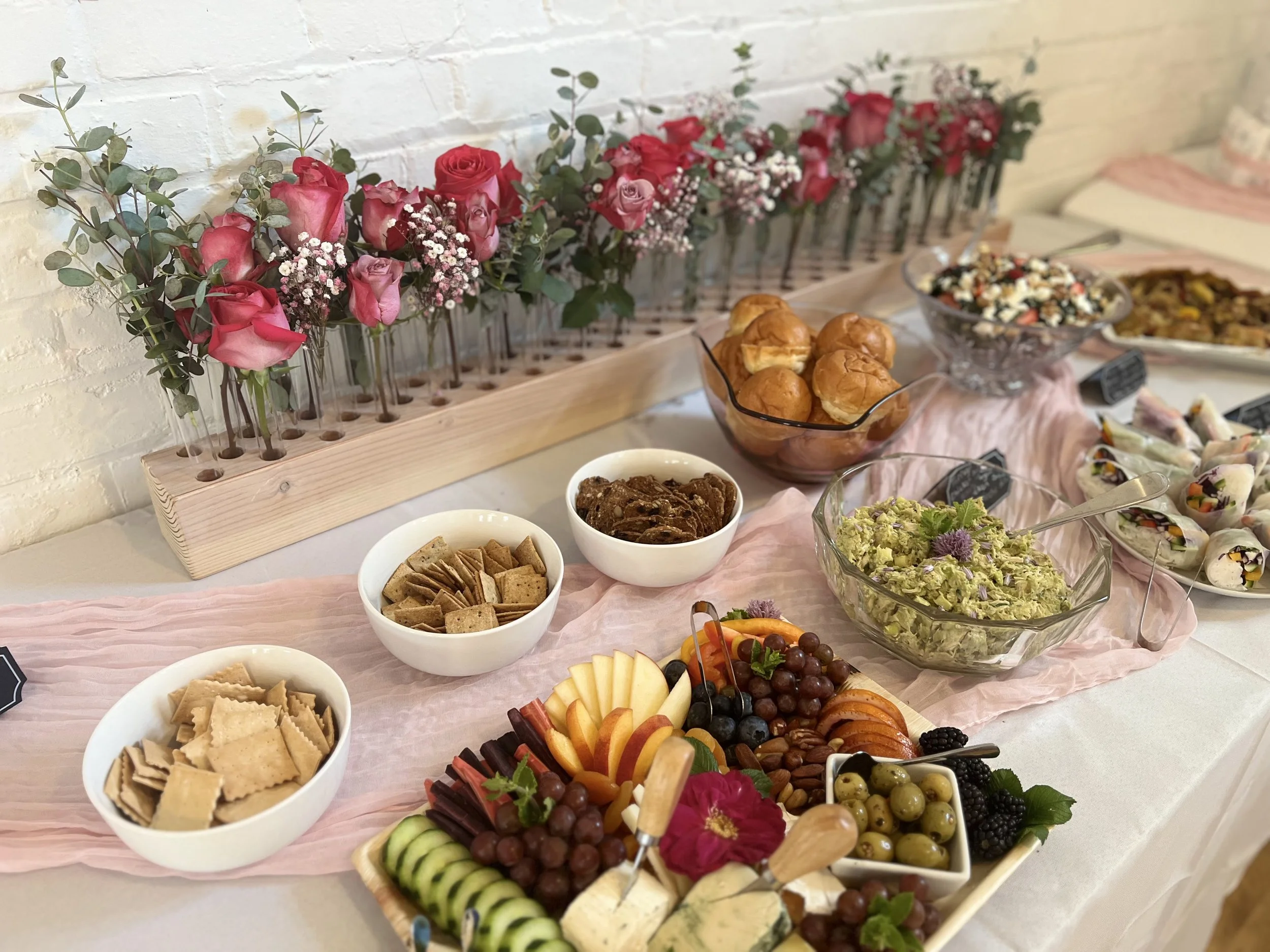 A tray with an assortment of sliced fruits, cheeses, grapes, and crackers on a table with pink fabric and floral decorations. Behind the tray are bowls of salad, chips, and snacks, with rose and eucalyptus decorations and a white brick wall in the ba