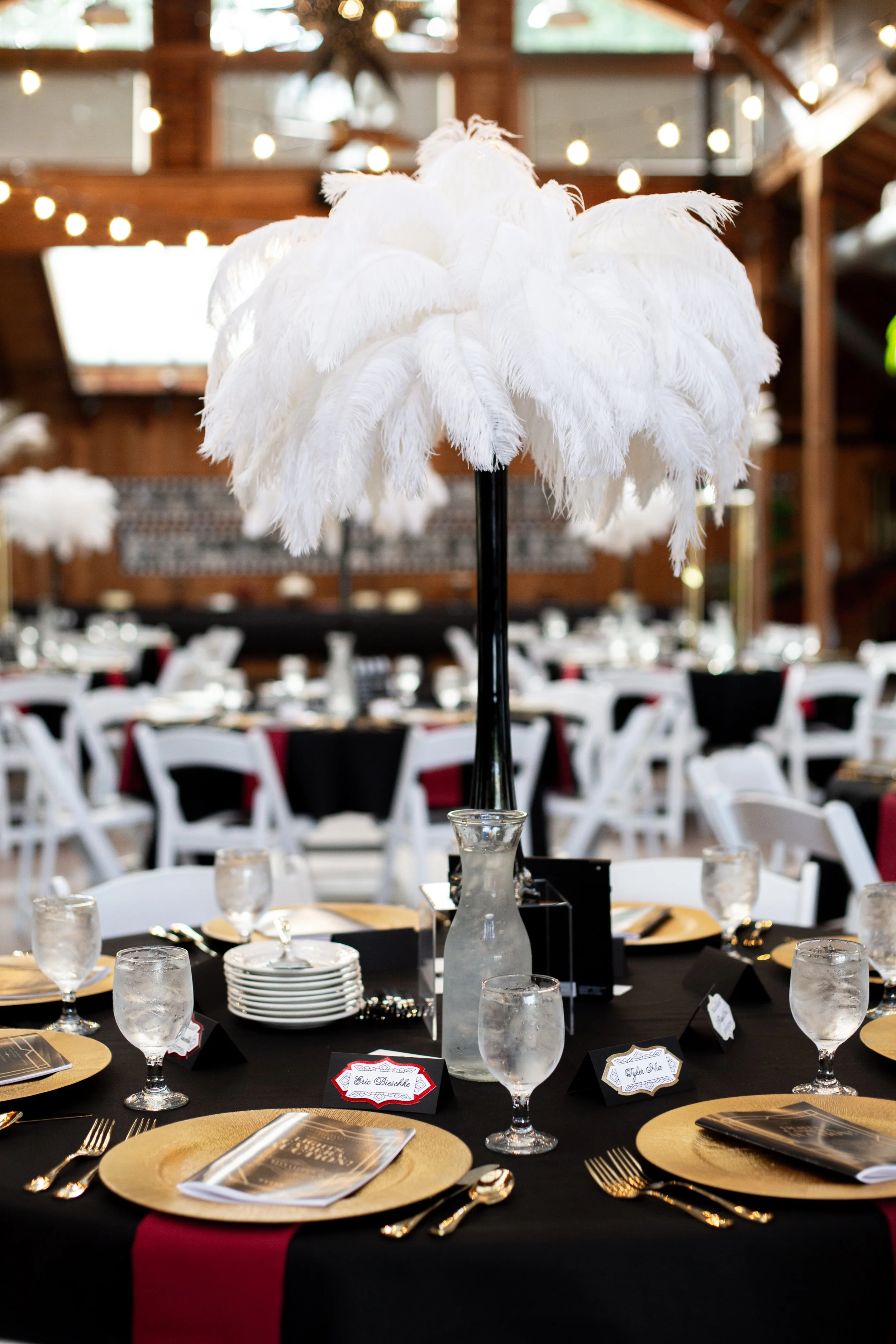 Elegant banquet table decorated with a tall centerpiece featuring white feathers, gold chargers, black tablecloth, gold cutlery, and water glasses, set in a rustic wooden venue with string lights overhead.