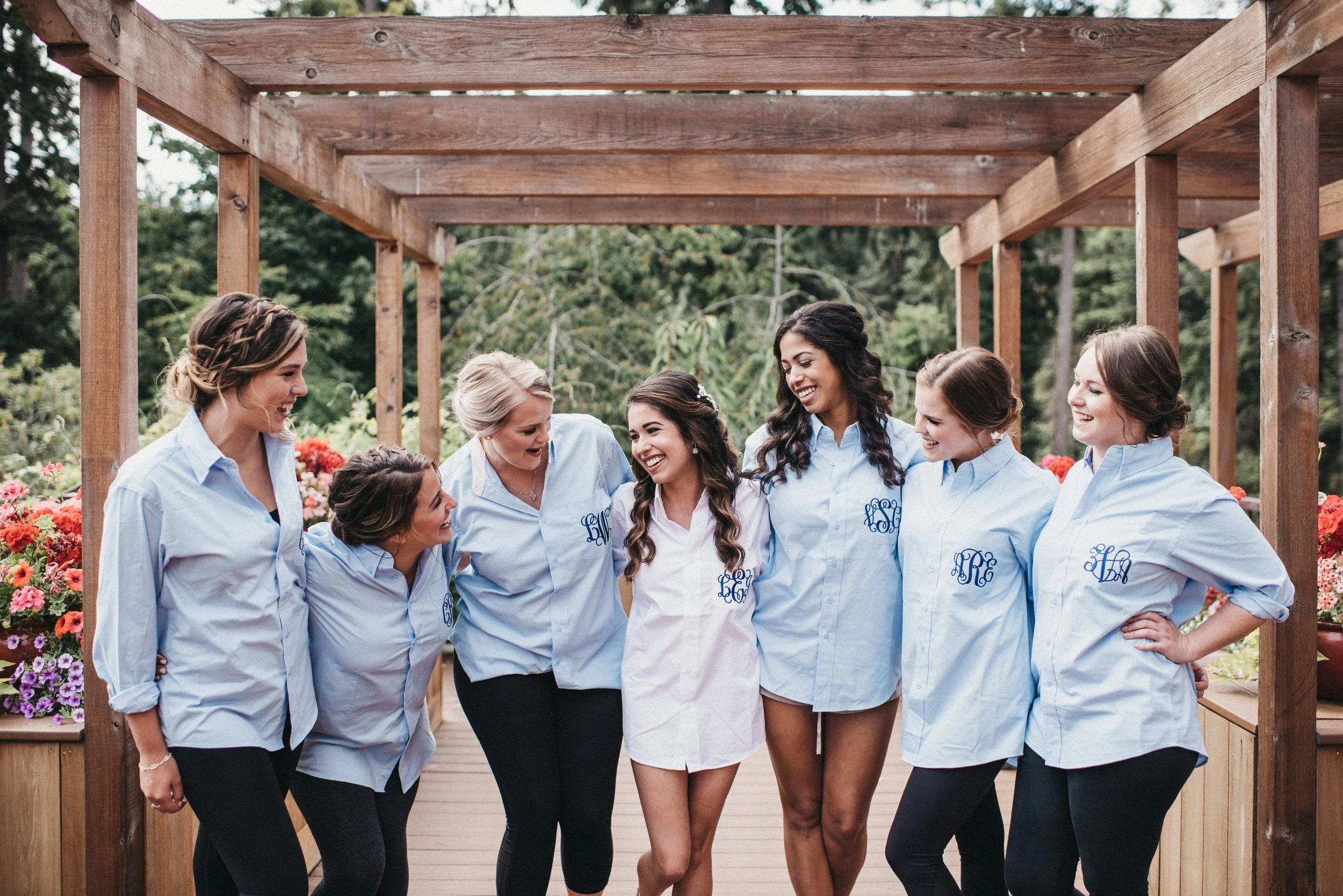 Group of seven women, one in a white dress and the others in light blue shirts with monograms, standing together on a wooden outdoor deck, smiling and enjoying each other's company.