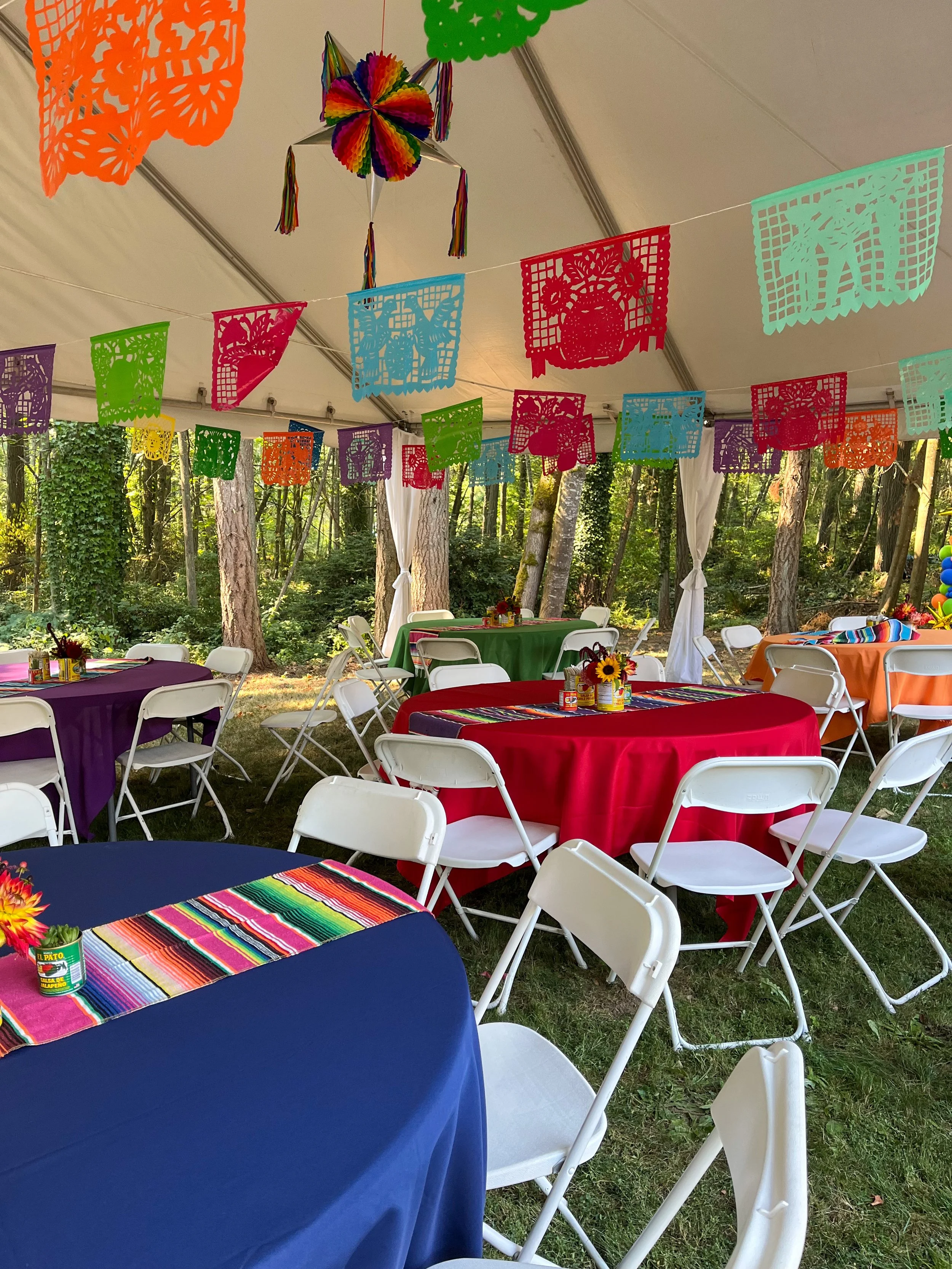 Colorful Mexican-themed party setup inside a tent with tables covered in bright tablecloths, white chairs, paper cut-out decorations hanging from the ceiling, and nature visible in the background.