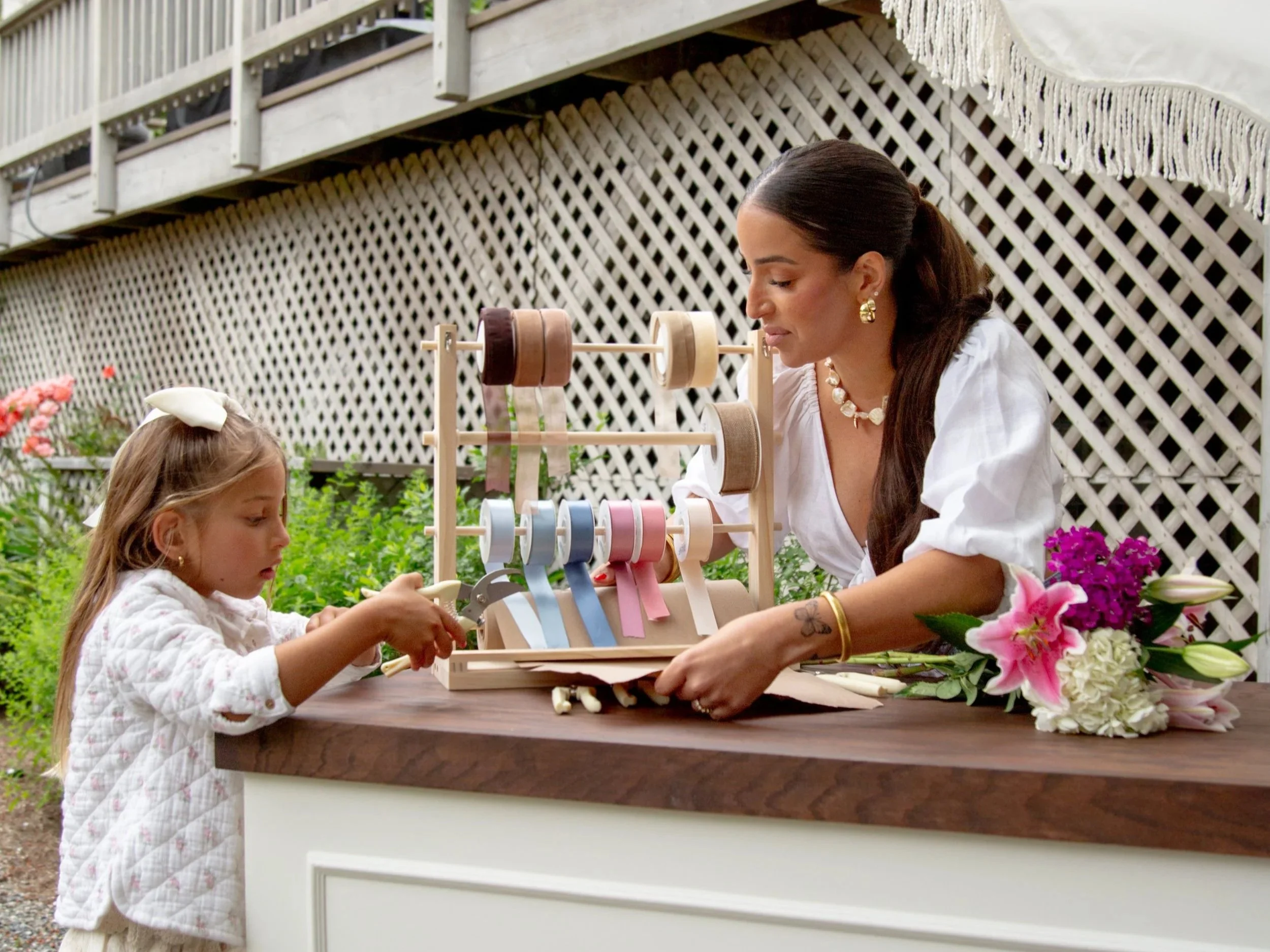 A woman and a young girl are working on a flower arrangement outside, with a wooden lattice fence and a deck with railing in the background. There is a flower display on the table.