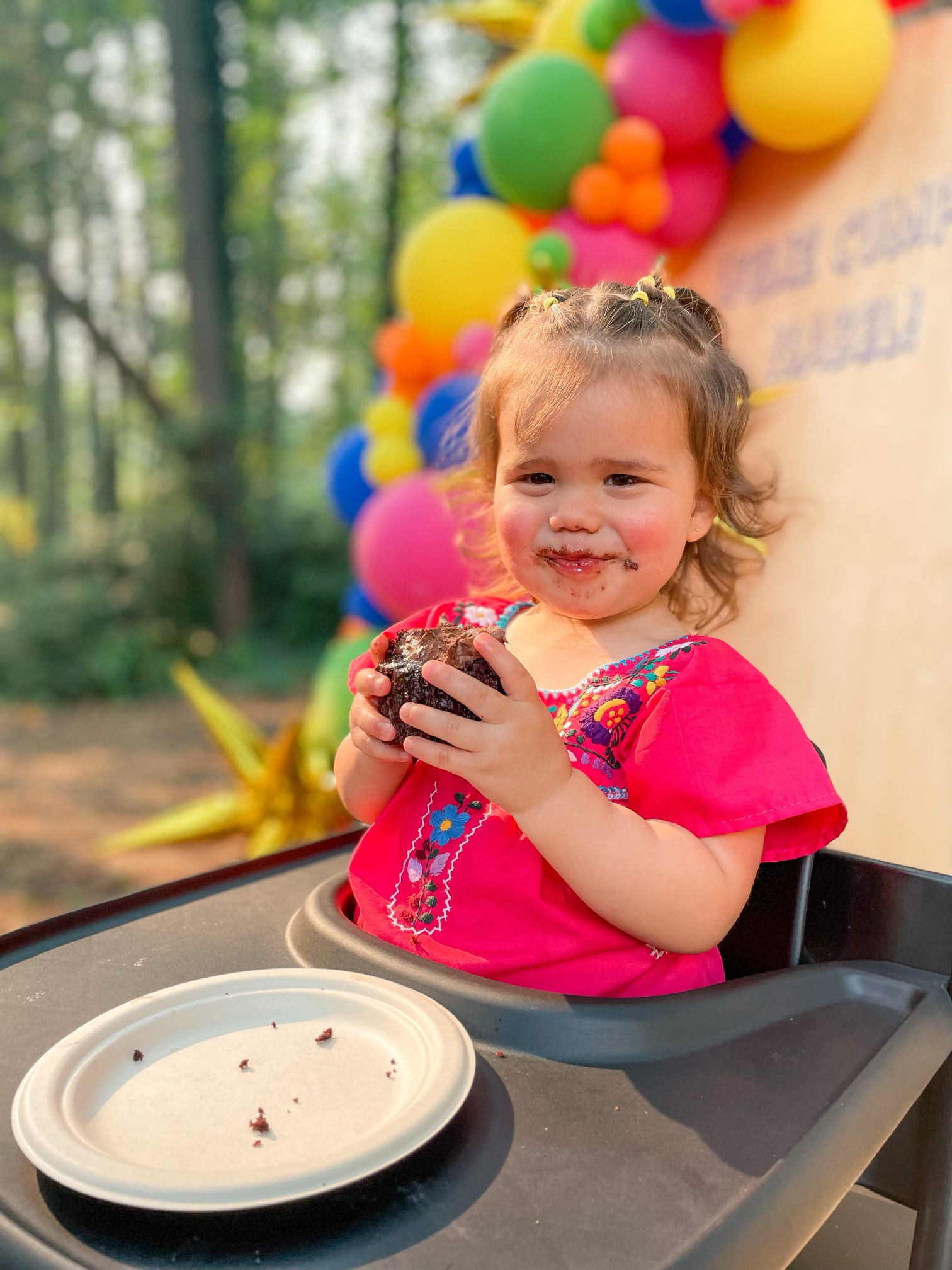 A young girl with curly hair and a pink embroidered blouse sitting in a high chair outdoors, holding a half-eaten chocolate cupcake, with a messy face and a slight smile. Behind her are colorful balloons.