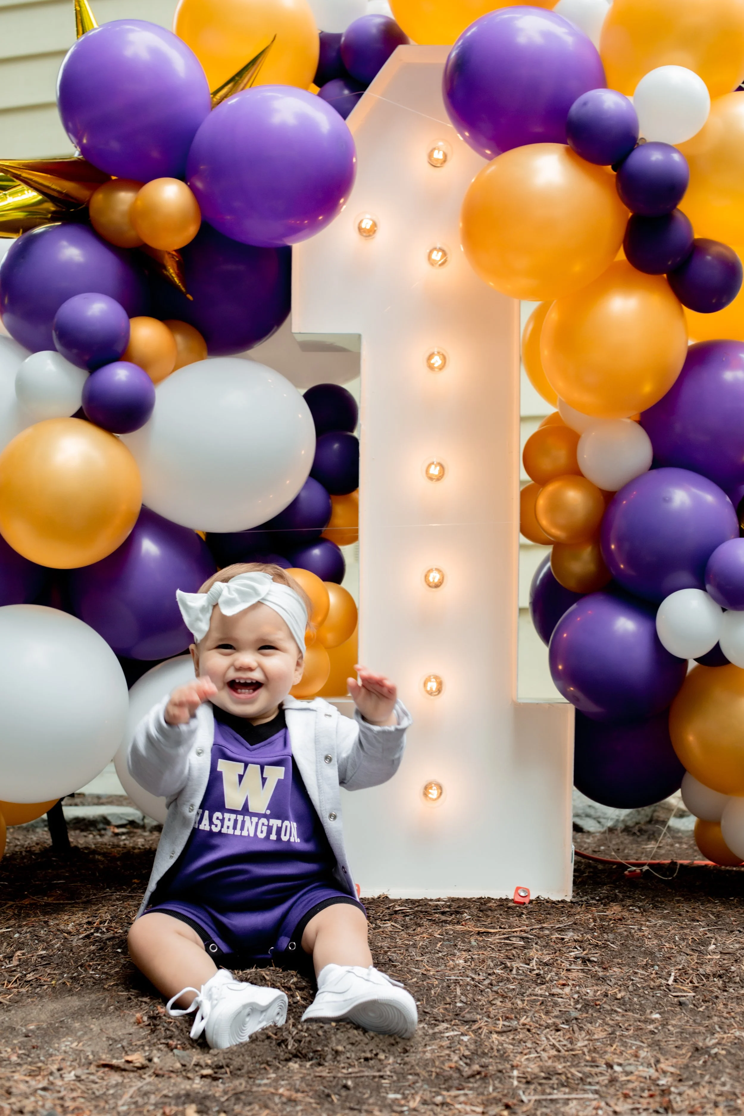 A young child sitting on the ground smiling, wearing a purple Washington jersey, white headband, white sneakers, and a light jacket, in front of a large illuminated number one surrounded by purple, gold, and white balloons, celebrating a first birthd
