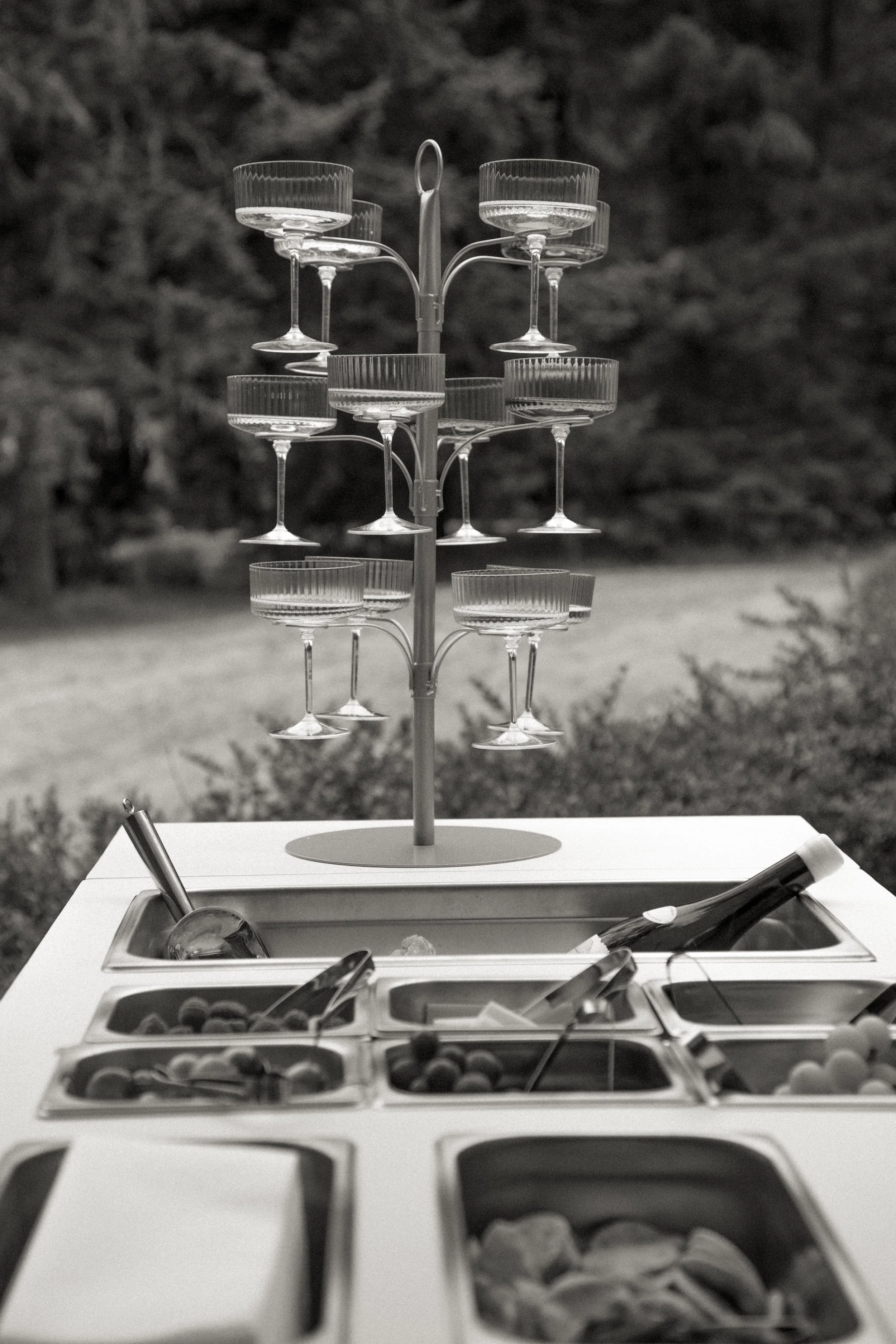 A tiered glassware stand with empty coupe glasses on a table, with a buffet of food in front, and a blurry outdoor background.