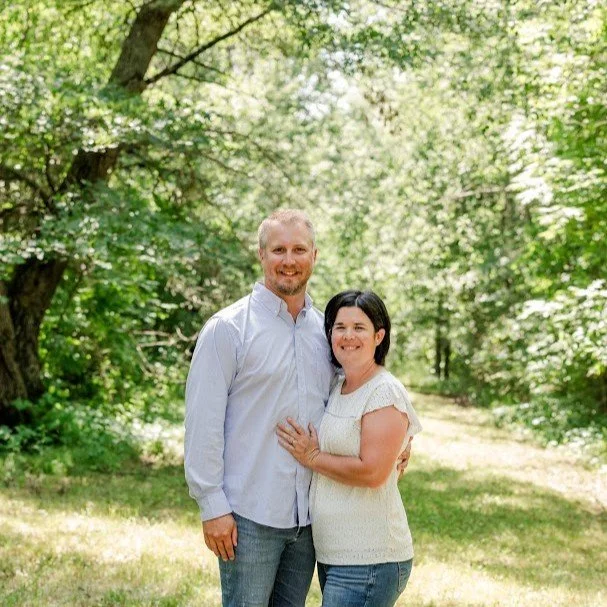 A couple embracing on a rural dirt road with greenery in the background, smiling and dressed casually.