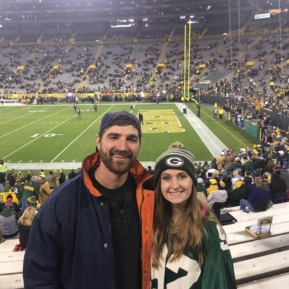 Two people are posing in the stands at a football stadium. They are wearing clothing with a Green Bay Packers logo. The field and end zone are visible in the background, with several people in the stands.