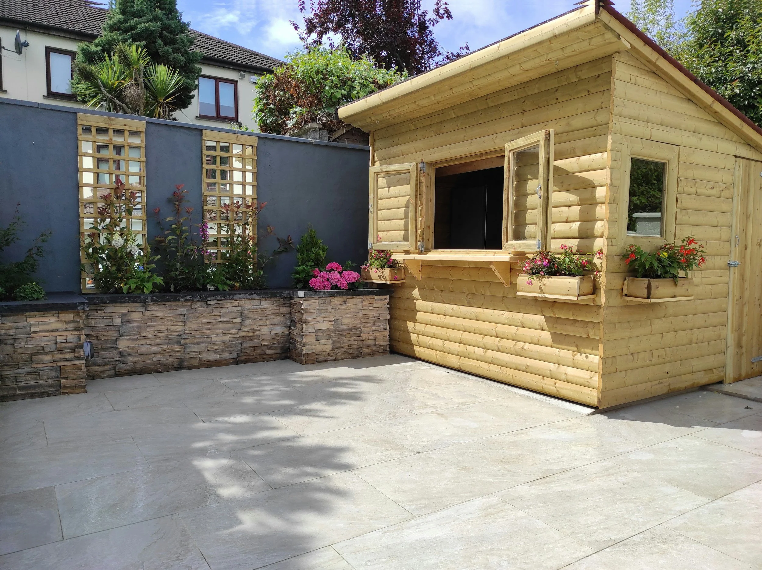 A small wooden garden shed with open window shutters and flower boxes, set on a patio with potted plants and a stone wall in a backyard.