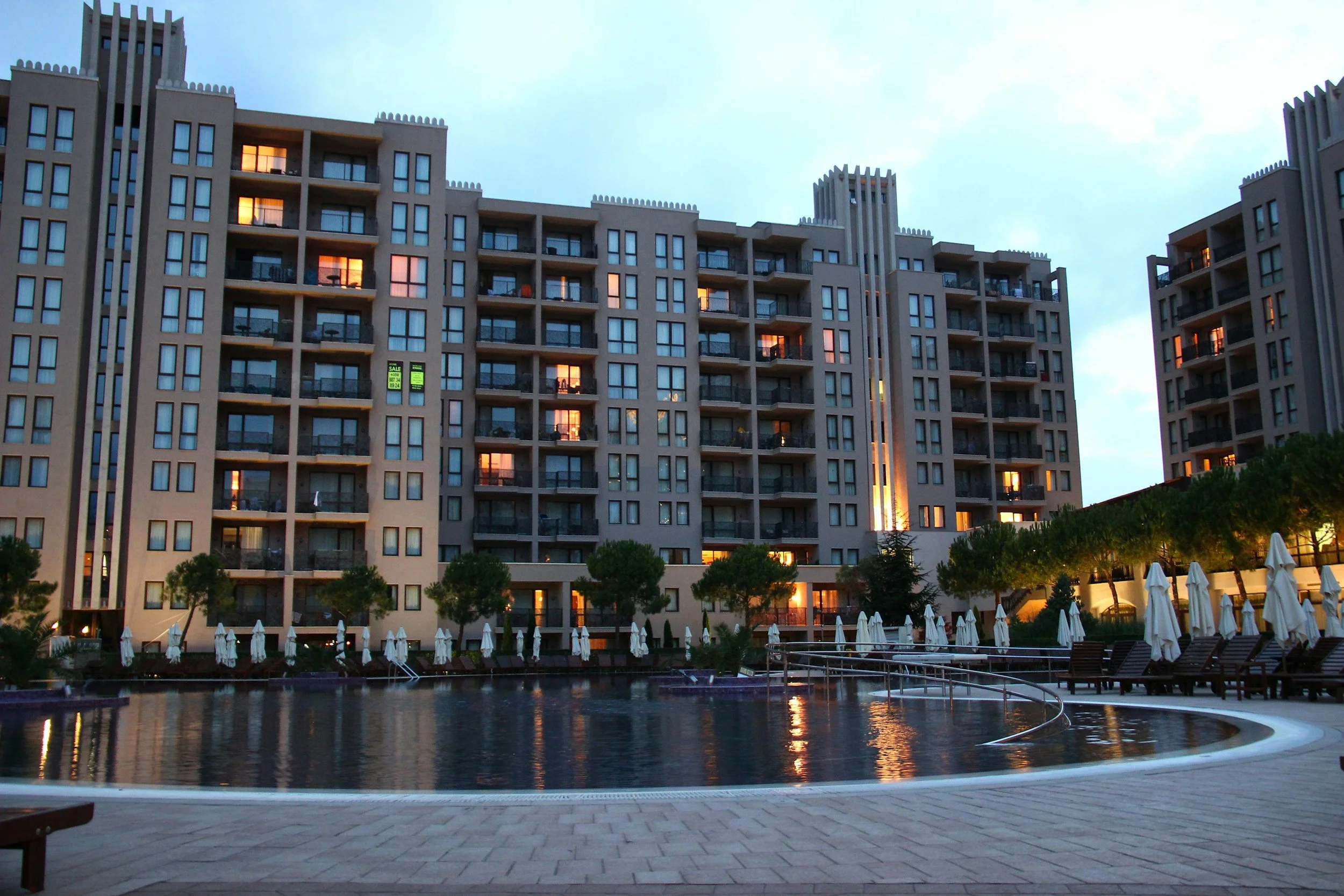 Modern apartment complex with a swimming pool, surrounded by umbrellas and trees, during twilight.