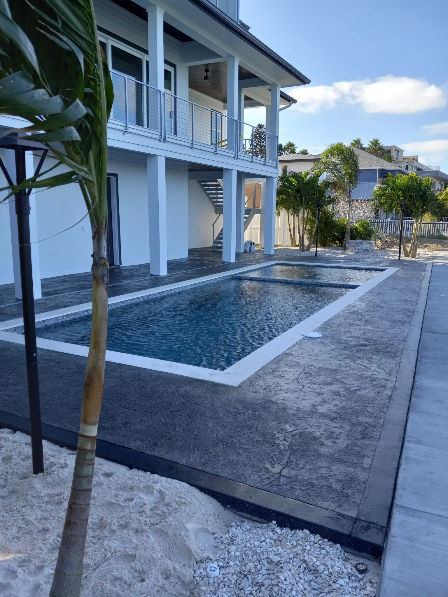 Modern house exterior with two swimming pools and a patio, surrounded by palm trees and neighboring houses, under a clear blue sky.